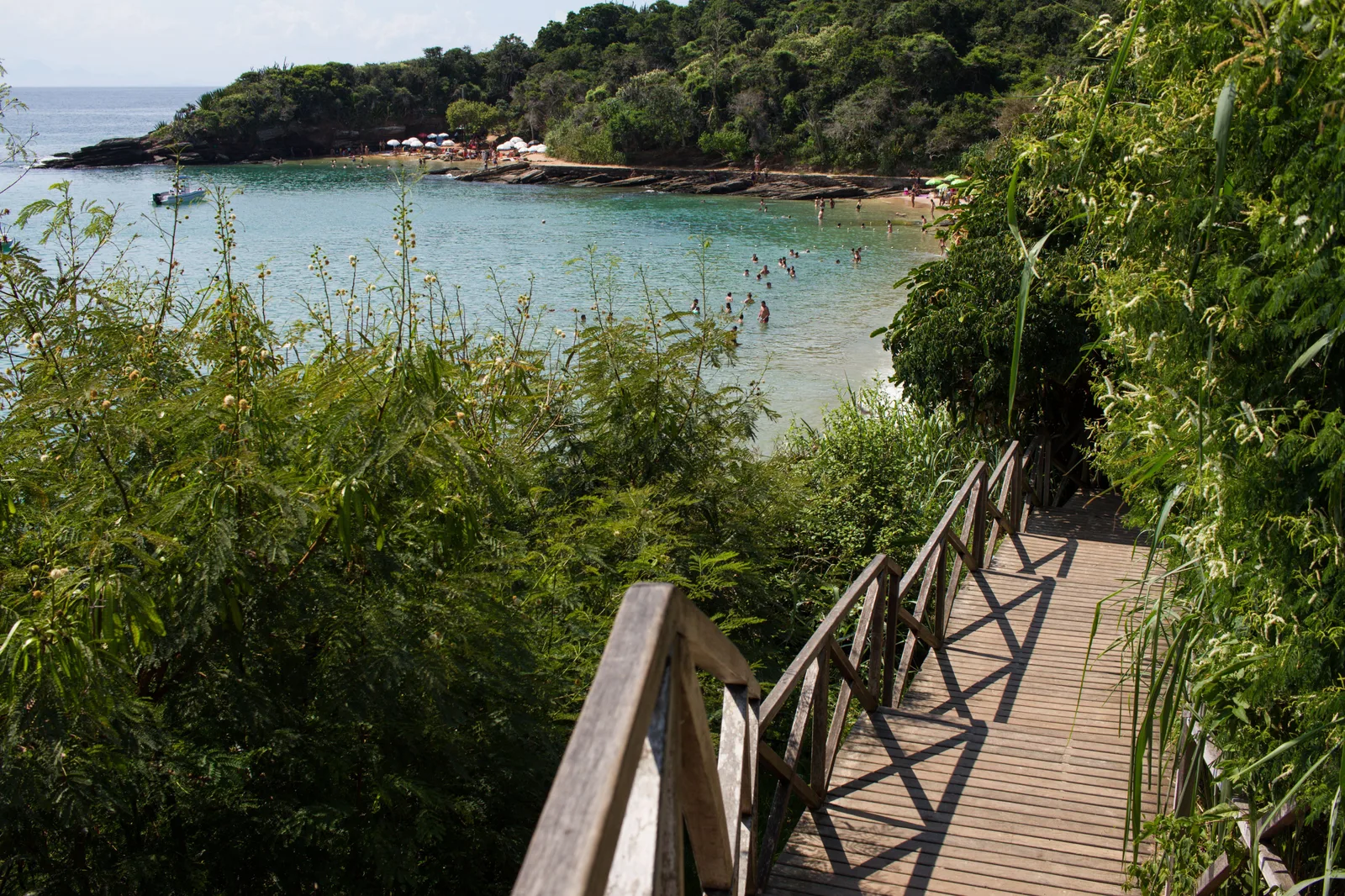 A wooden stairway with wooden railings running down through green scrub toward a small cove at Búzios, with swimmers in clear turquoise water and parasols on the sand beyond
