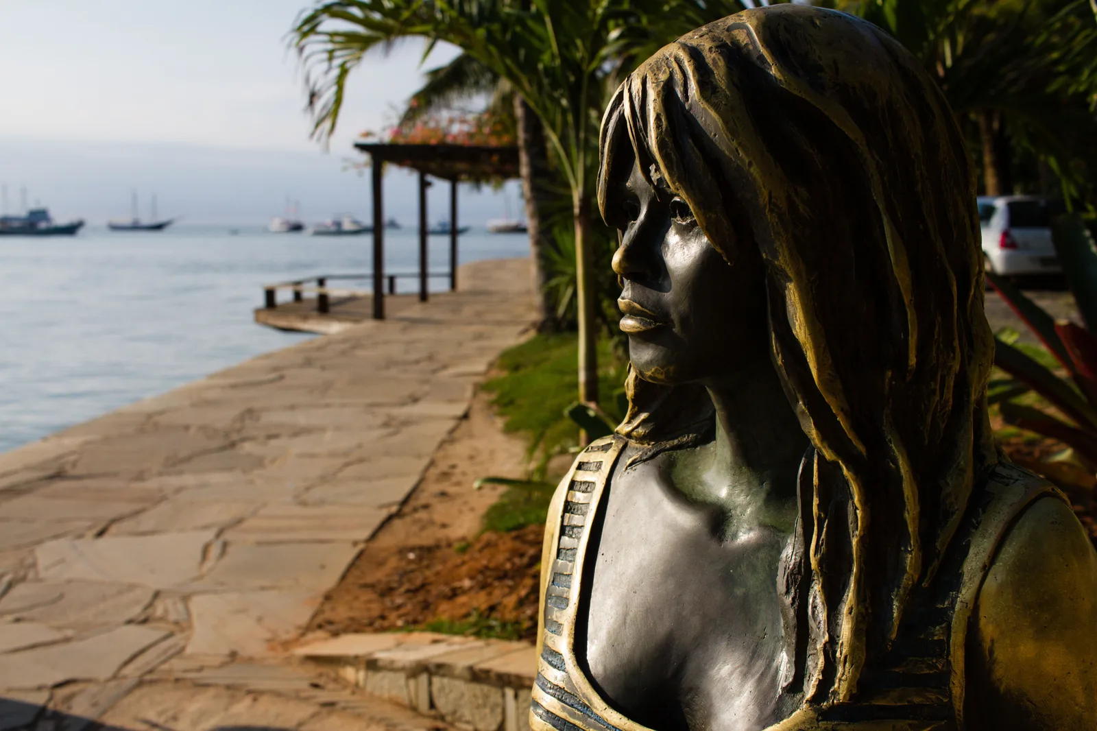 The head and shoulders of the bronze Brigitte Bardot statue on the Orla Bardot at Búzios, close up in profile, with the paved waterfront walk, a pergola, palms and moored boats on the bay behind