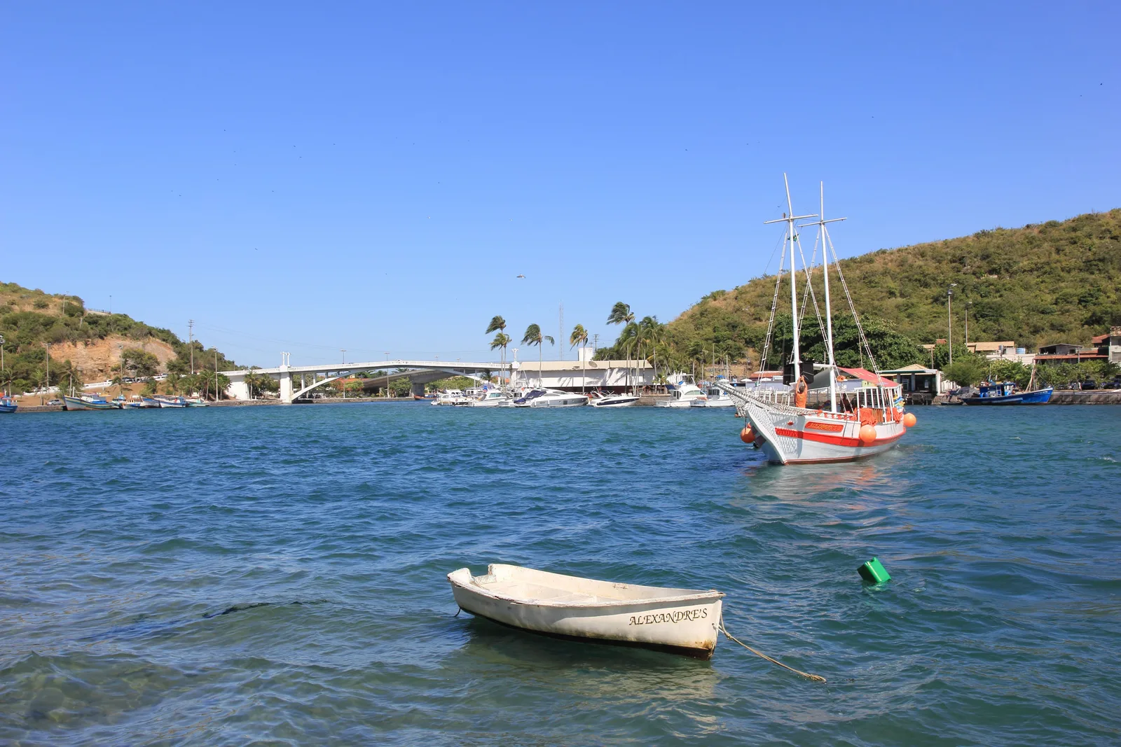 The Canal do Itajuru at Cabo Frio: a two-masted schooner and small boats on flat blue-green water, with a road bridge crossing the channel and a green hill behind