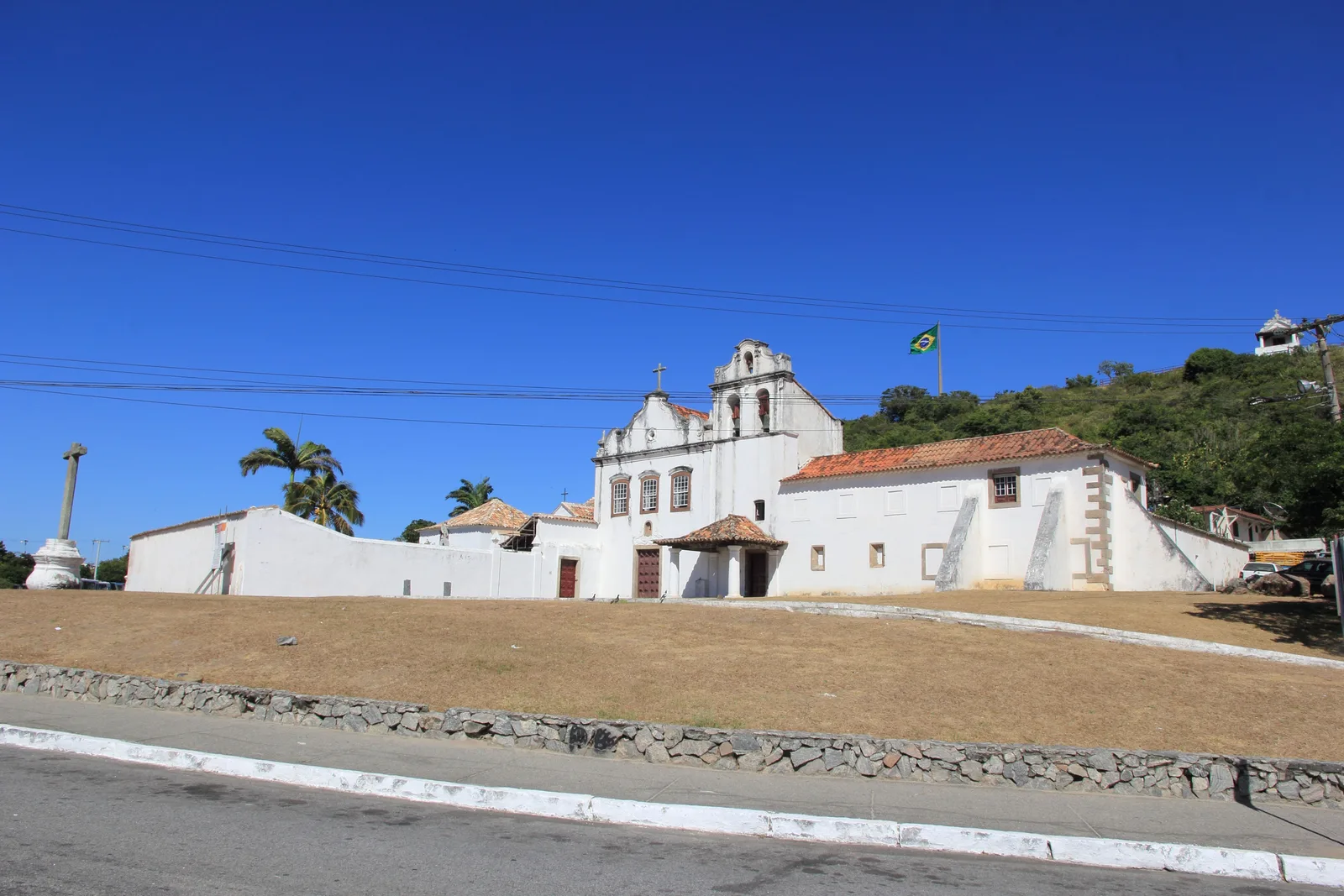 The white Convento de Nossa Senhora dos Anjos at Cabo Frio: a long low colonial building with tiled roofs and a curved bell gable, behind a sloping lawn of dry grass, with a stone cross on a column at one end and a green hill behind