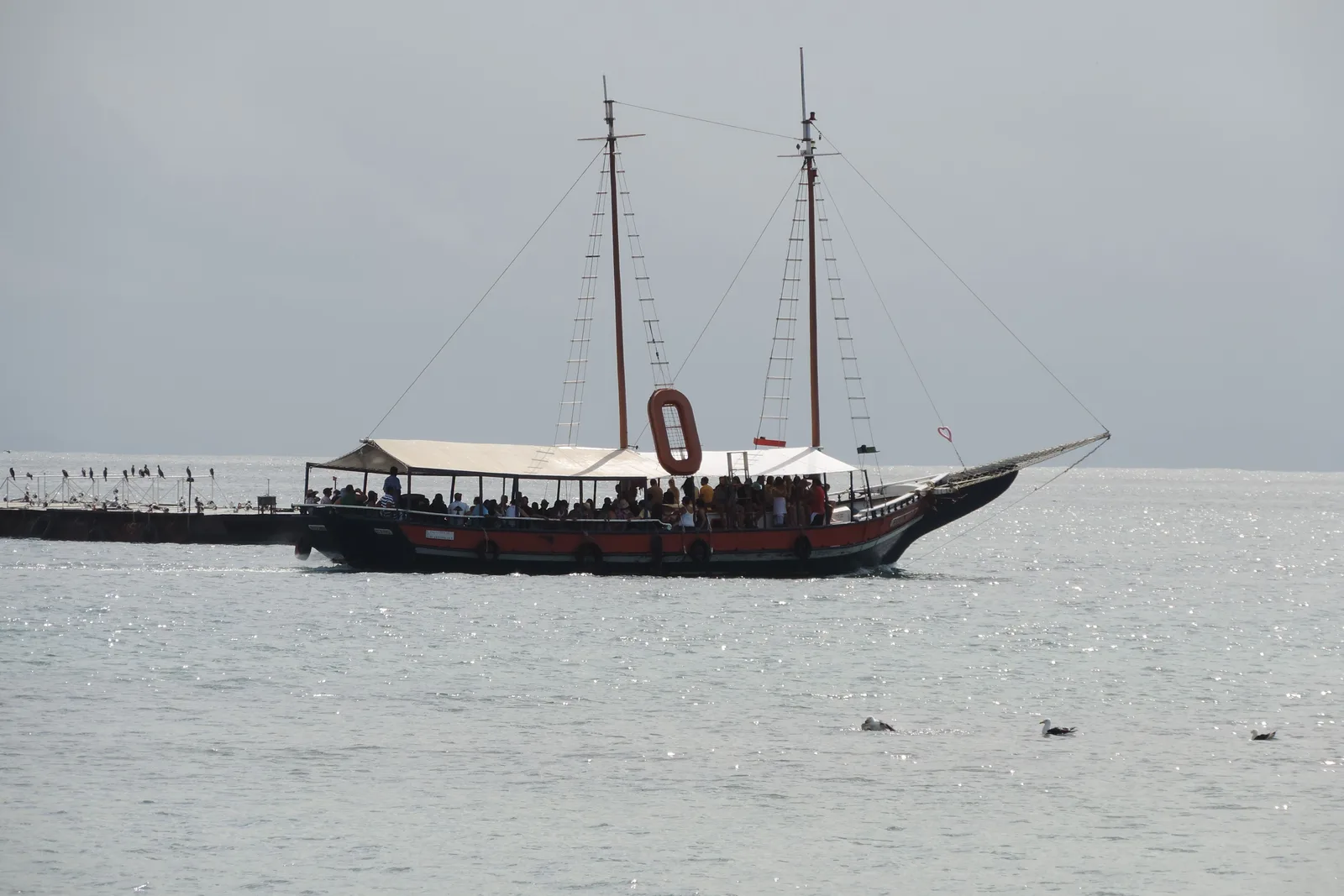 A two-masted wooden schooner with a red and black hull and a white canopy over a deck full of passengers, on silver-gray water in the bay at Búzios under an overcast sky, with a jetty behind it