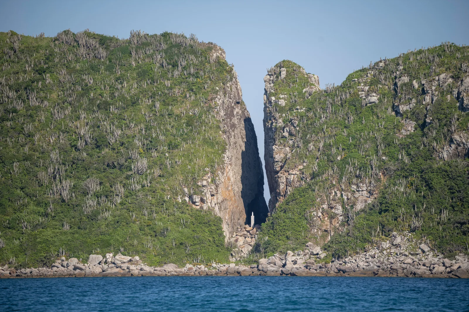 A narrow vertical cleft splitting a steep, cactus-dotted coastal cliff on Ilha do Cabo Frio from the ridge almost to the sea, with a small white statue standing on the boulders at its foot and a rocky shore along the waterline