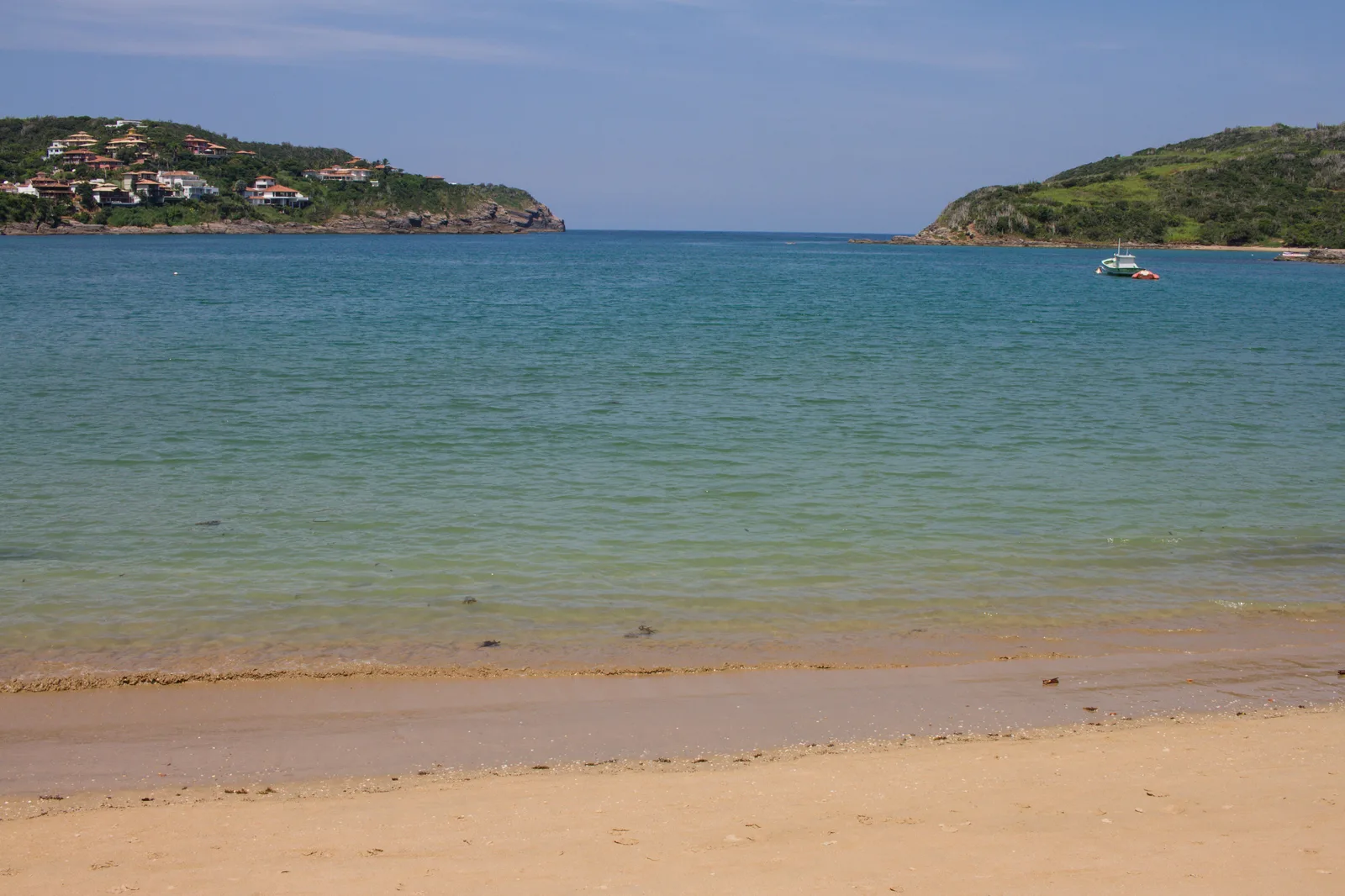 Calm pale blue water inside Praia da Ferradura in Búzios seen from the sand, with two green headlands framing the narrow mouth of the bay, houses on the left-hand one and a small boat at anchor