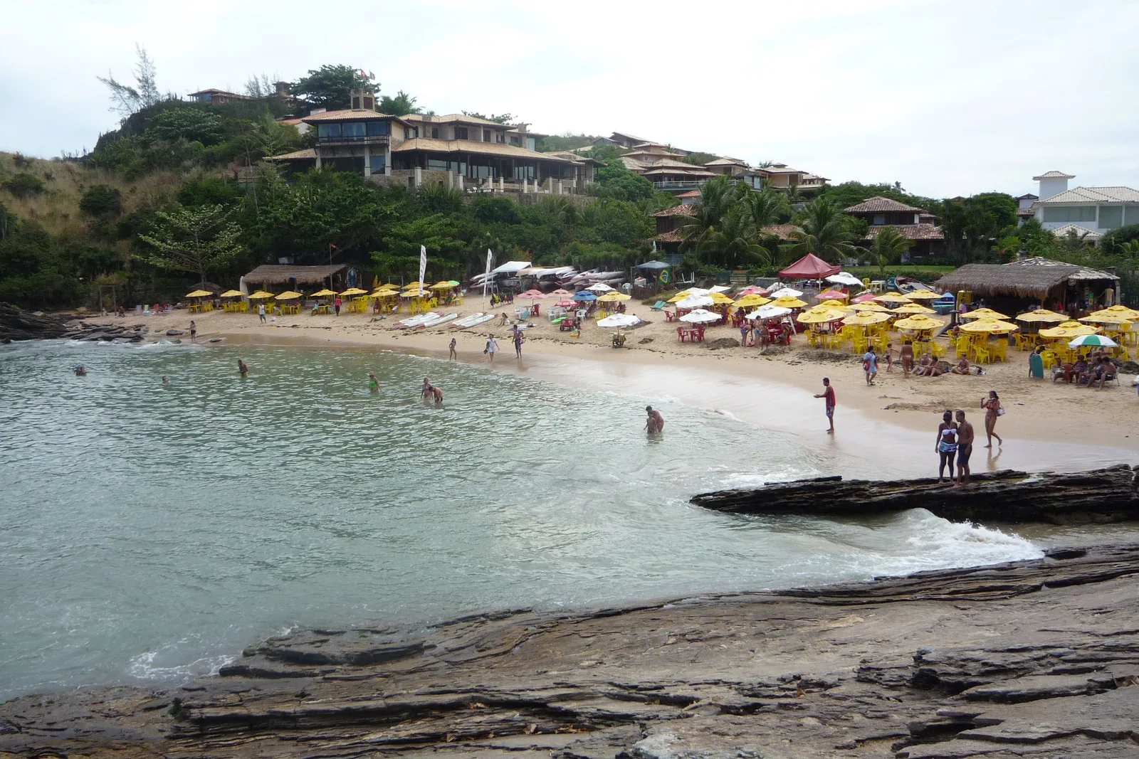 The small cove at Ferradurinha in Búzios seen from the flat rock shelf at one end: a crowded beach lined with yellow parasols and kiosks, swimmers in green water, and large houses on the wooded slope behind