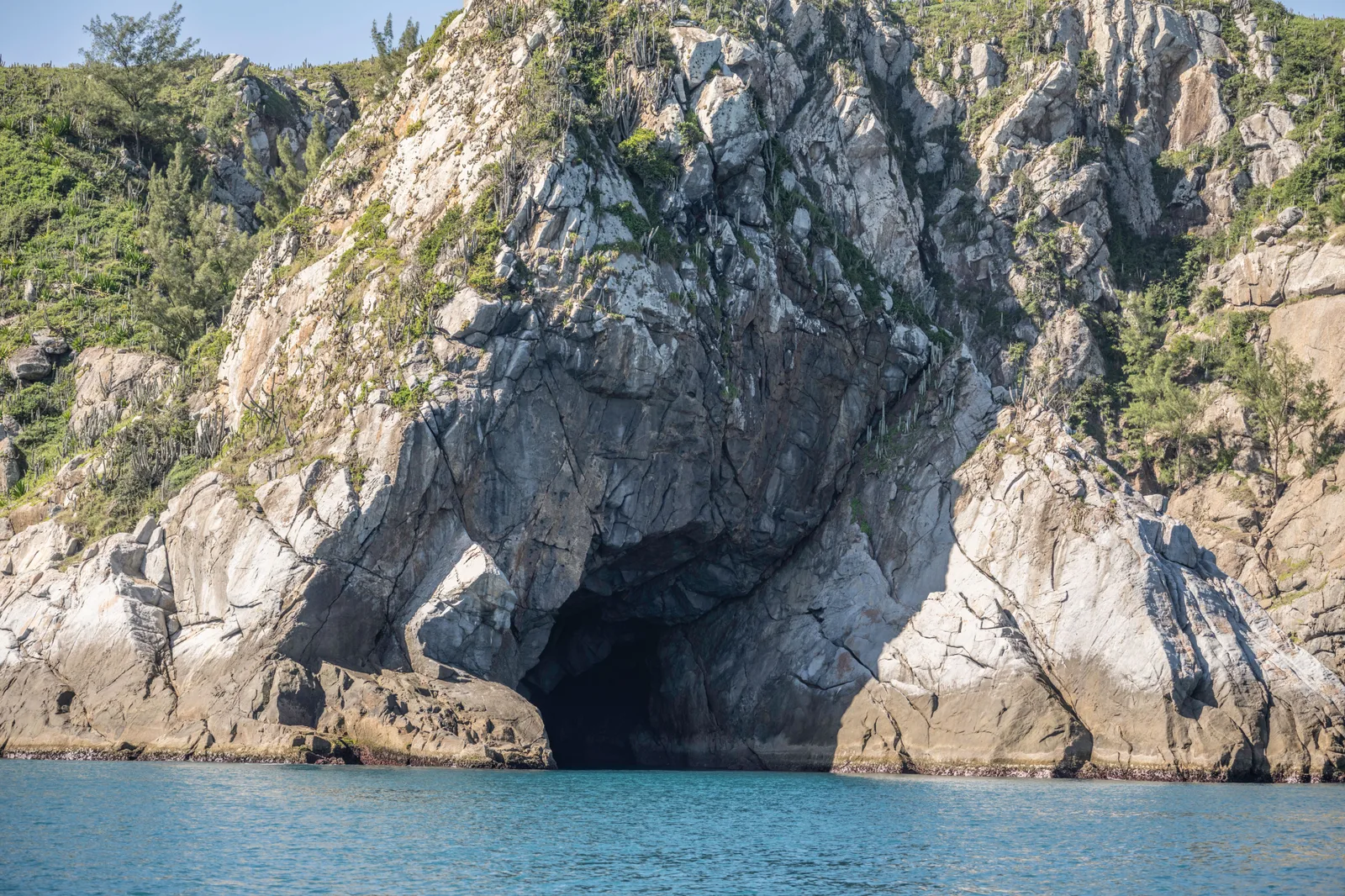 A pale gray rock headland on Ilha do Cabo Frio with a wide dark cave mouth at the waterline, green scrub growing over the rock above it and turquoise water washing in