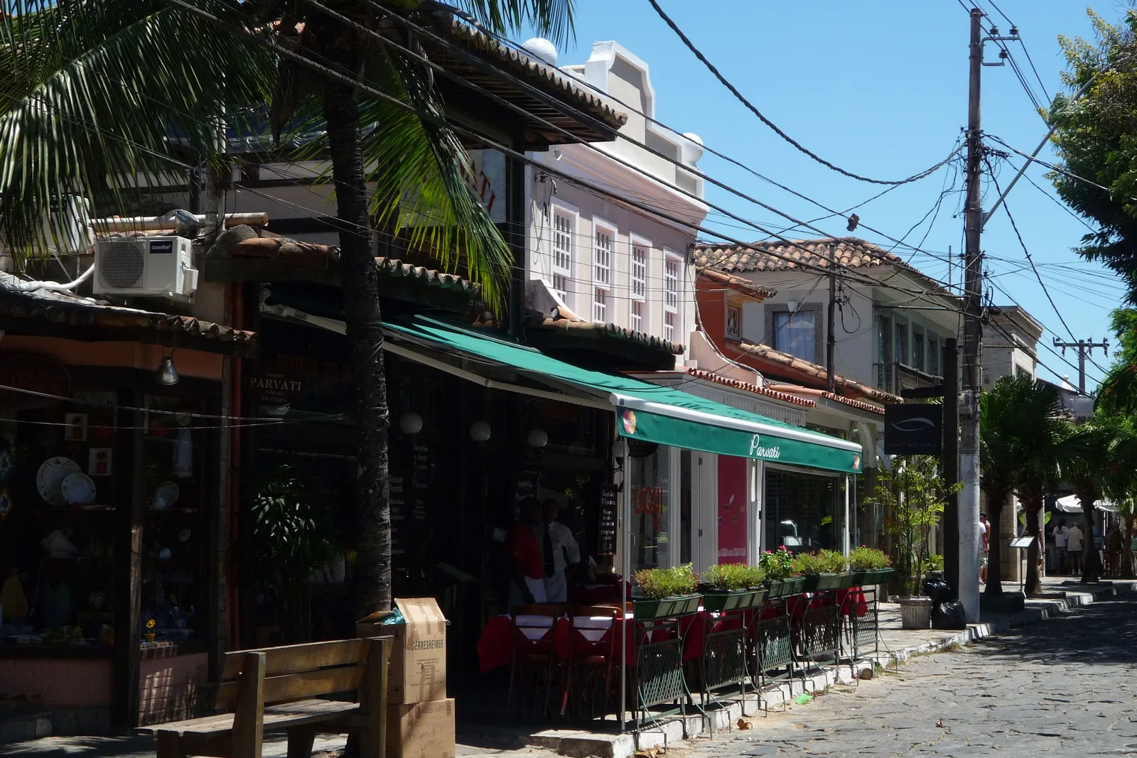 A restaurant terrace under a green awning on the stone-paved Rua das Pedras in Búzios, with tables in red cloths set out behind a railing of planters
