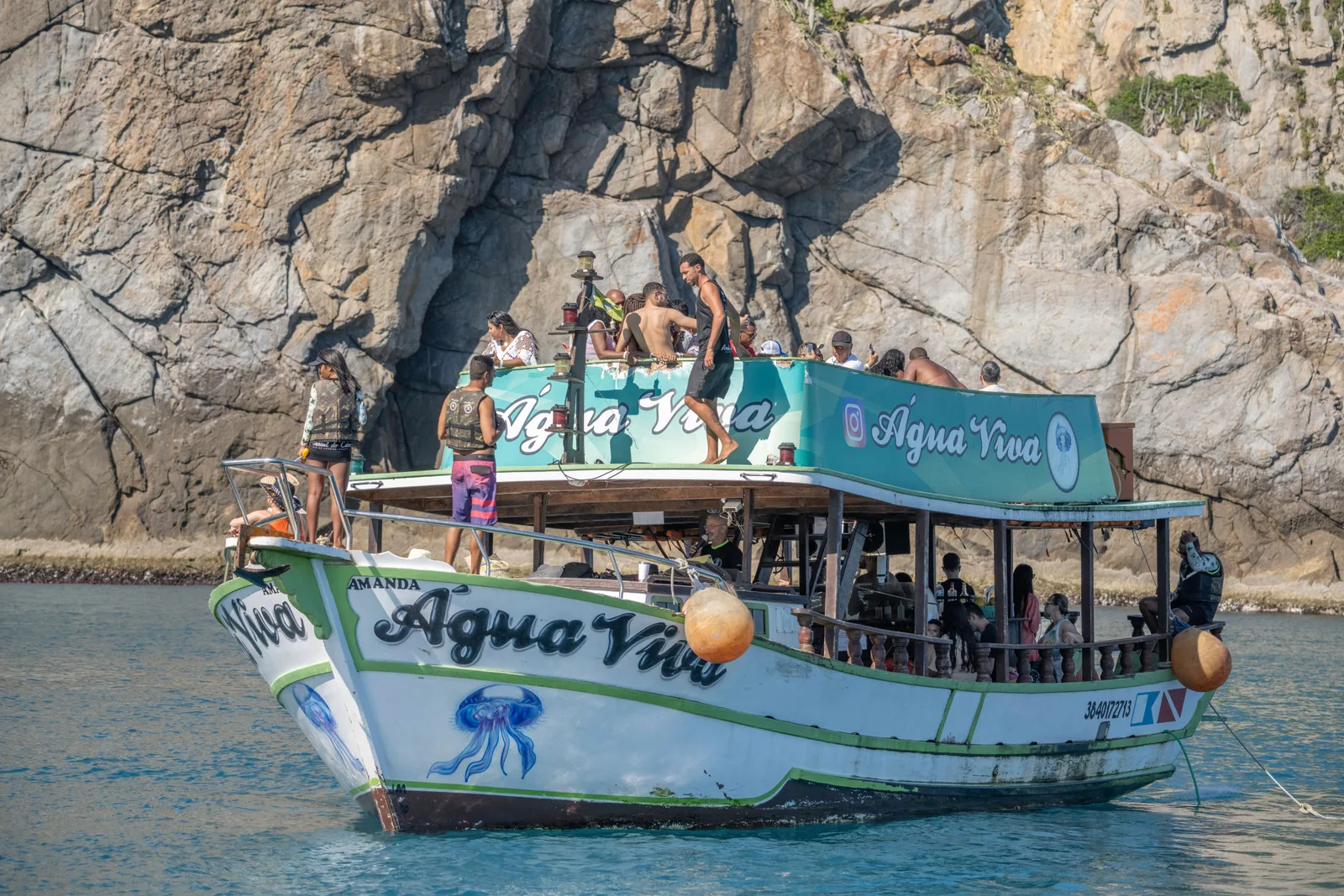 A wooden tour boat painted white and green, with passengers on both decks and a jellyfish painted on its bow, anchored in front of a tall gray rock cliff at Arraial do Cabo