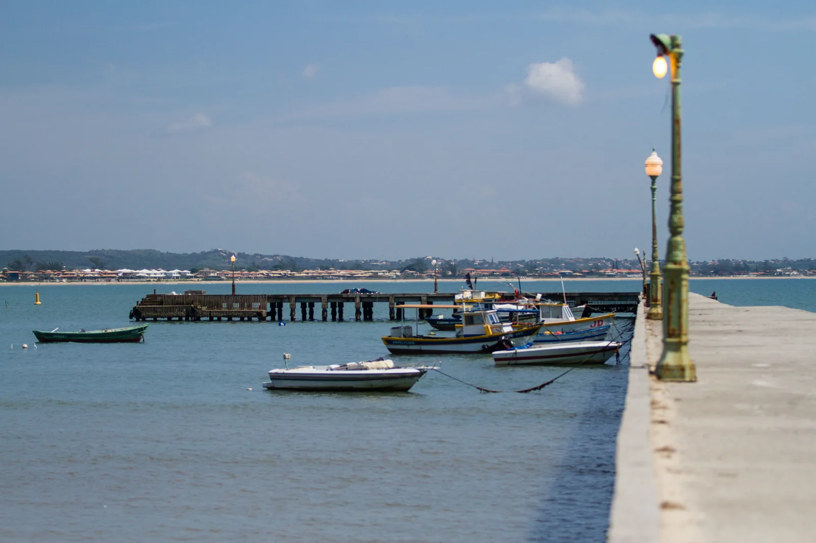 A concrete pier at the Porto da Barra in Búzios with green lamp posts along it, a long wooden jetty across the water, and small fishing boats moored on the flat water beside it