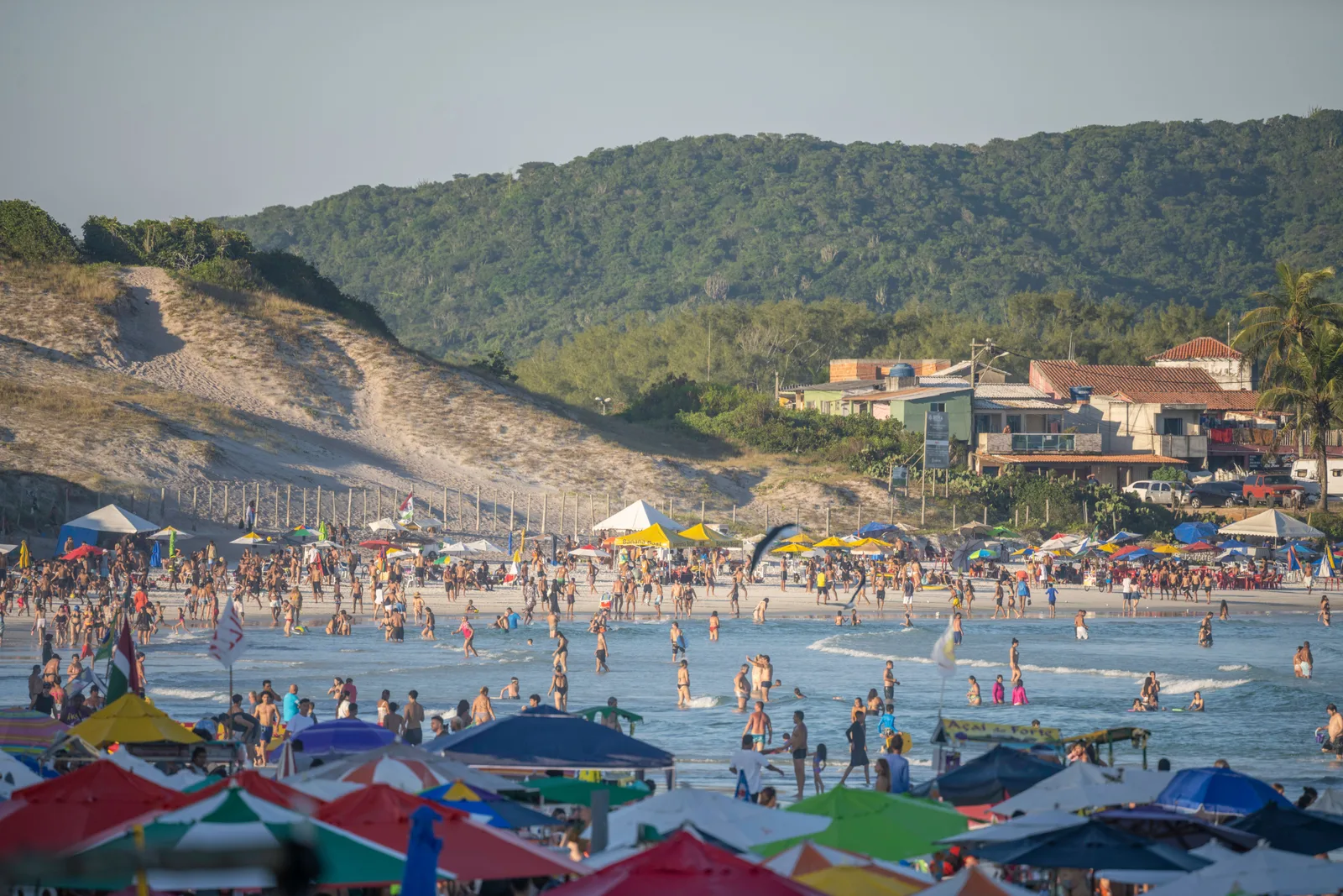 A crowded stretch of beach at Cabo Frio packed with colored parasols and swimmers, with white sand dunes and a wooded green hill rising behind the far end