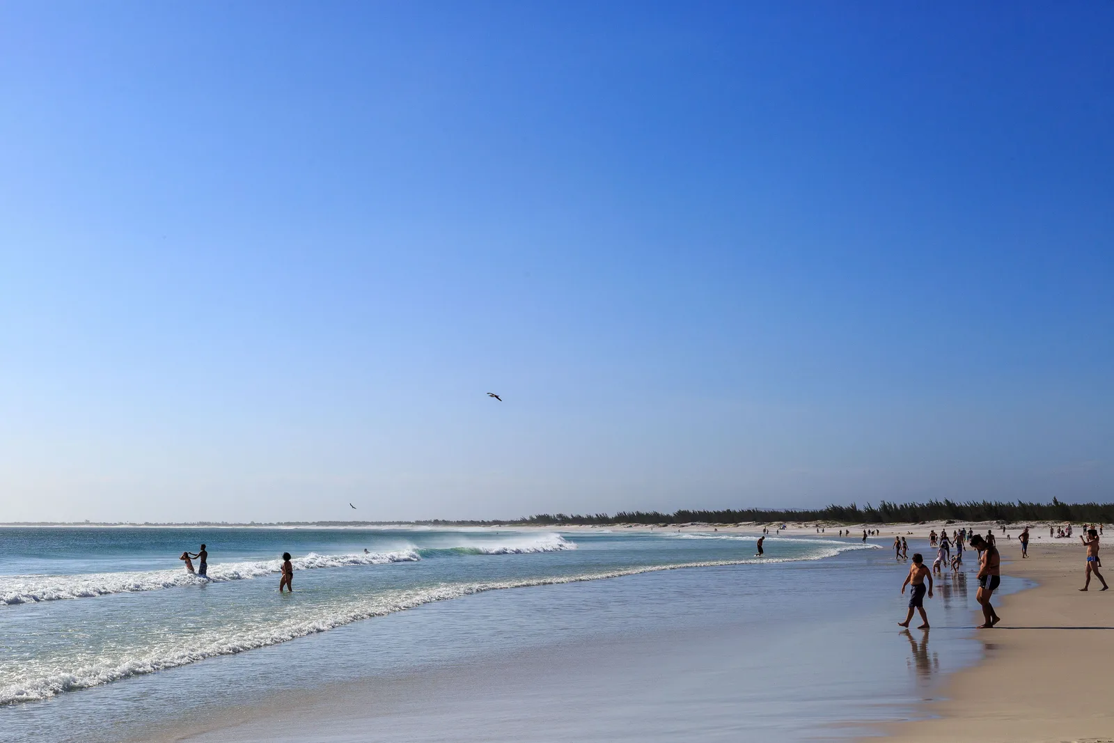 The long open beach at Praia Grande in Arraial do Cabo with white surf running up the wet sand and small groups of people walking along the waterline