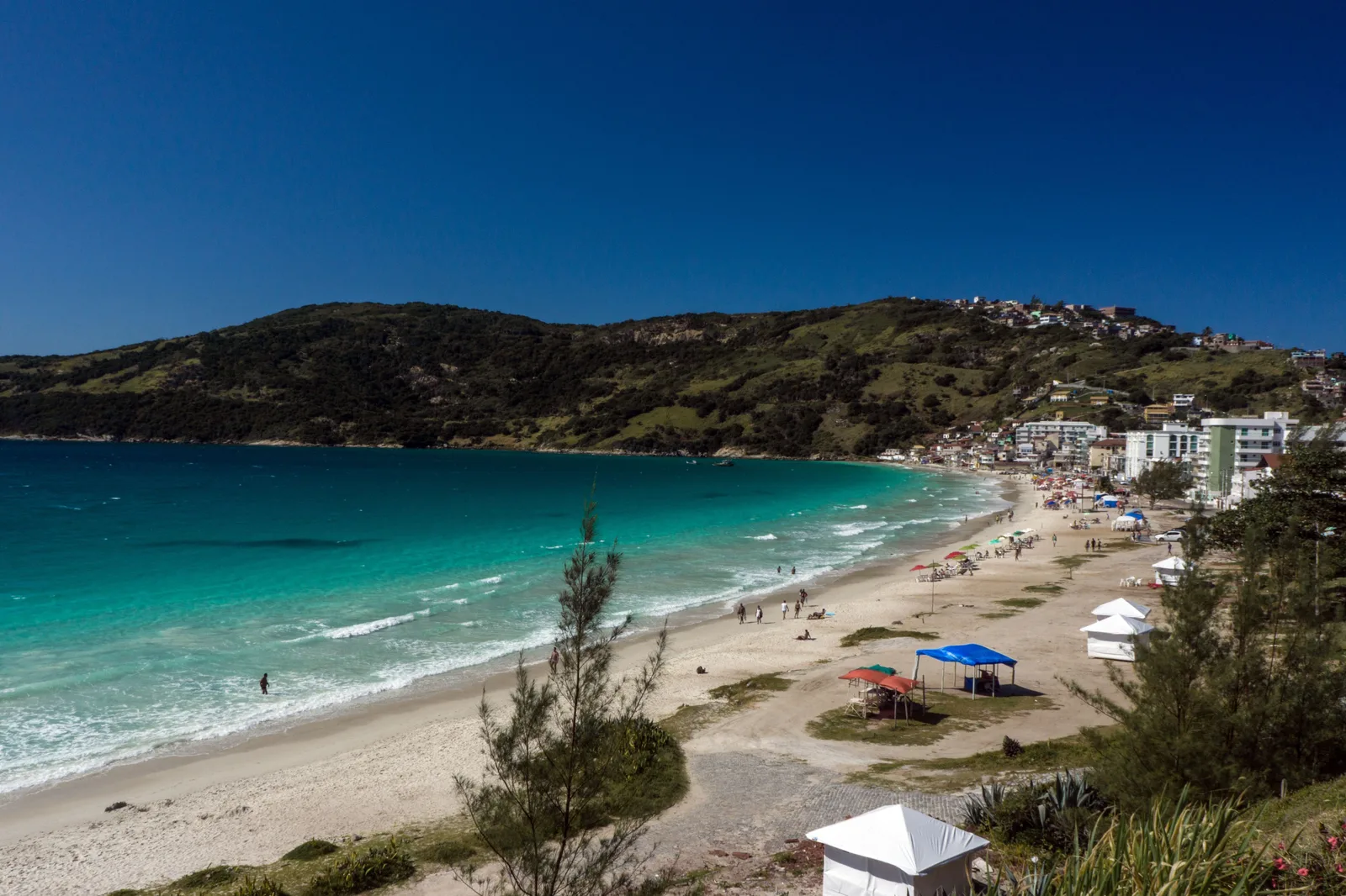 The Prainha at Arraial do Cabo seen from a low rise behind the beach: a curve of sand with sunshades and small tents along it, turquoise water with small waves breaking, apartment buildings at the town end and a green headland behind