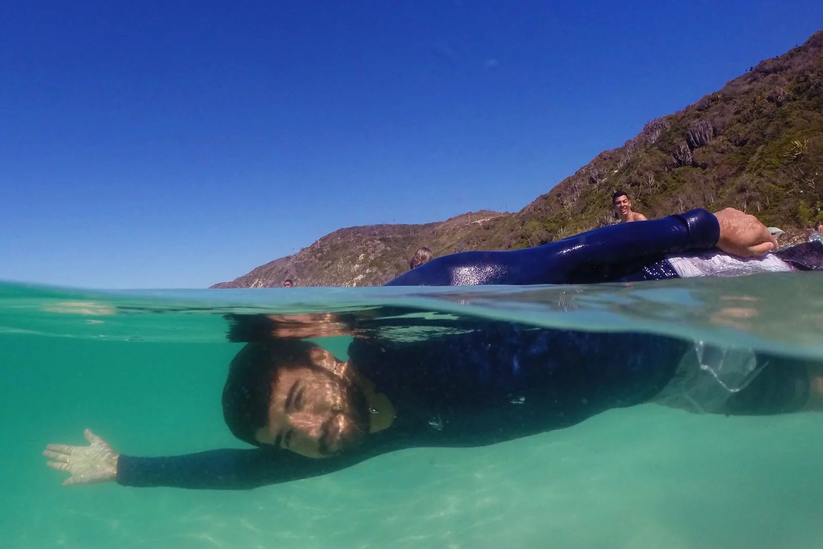 A half-underwater photograph at Arraial do Cabo: a person in a blue wetsuit floating at the surface with their face visible through clear green water, and a green headland and blue sky above the waterline