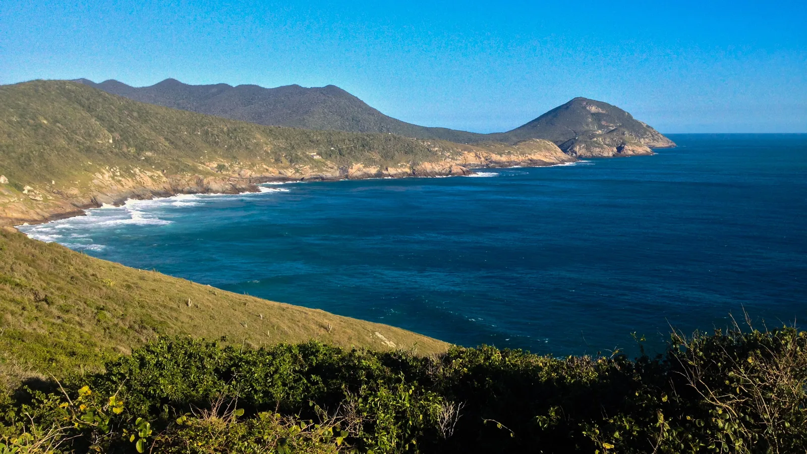 The coastline of the Costa do Sol from a green hillside: a long curve of dark blue sea against steep scrub-covered headlands running away toward a distant point