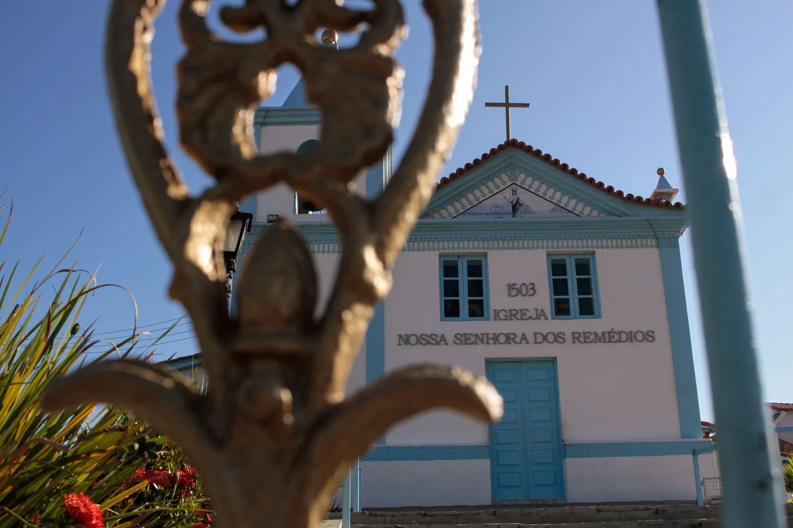 The white and blue facade of the Igreja Nossa Senhora dos Remédios in Arraial do Cabo, with 1503 and the church’s name lettered above its blue door, seen past a blurred bronze railing finial in the foreground
