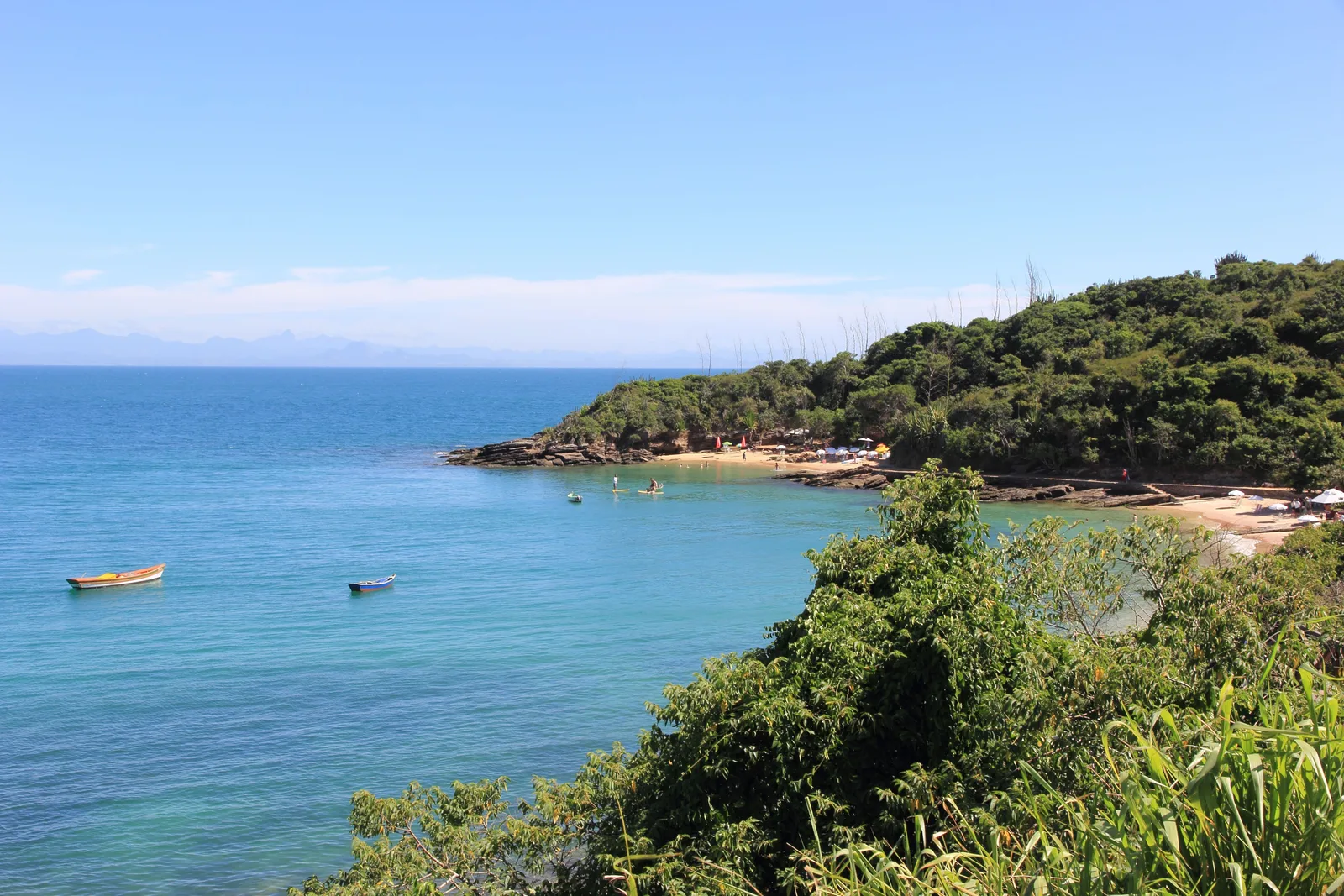A small Búzios cove seen from the hillside above: parasols along the sand at the foot of a wooded headland, clear blue water with two small boats on it, and green shrubs in the foreground