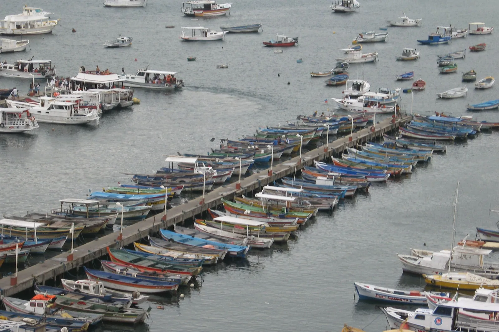 A long jetty at Praia dos Anjos in Arraial do Cabo seen from above, with dozens of small painted boats moored in rows along both sides of it and larger tour boats anchored beyond