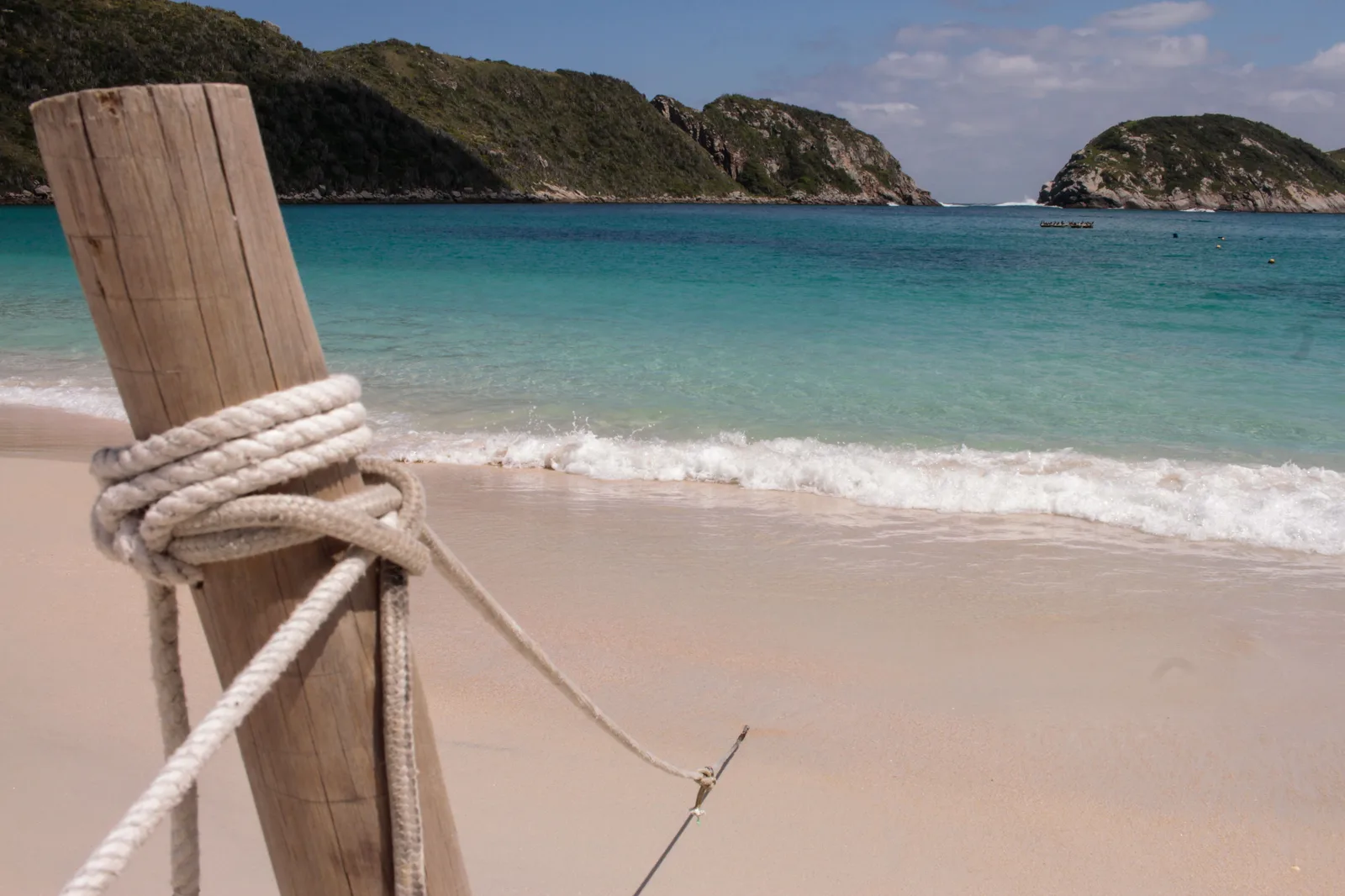 A rope knotted around a weathered wooden post on the sand on Ilha do Cabo Frio, with small waves breaking, clear turquoise water, and green rocky headlands and an islet behind it