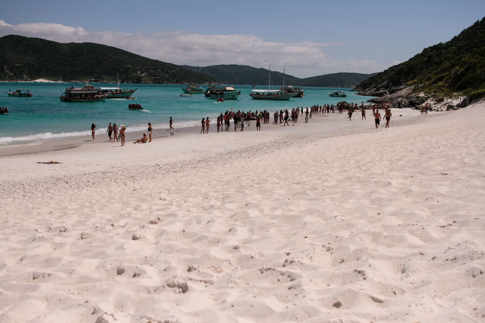 A crowd of people at the waterline of Praia do Farol on Ilha do Cabo Frio, with a wide stretch of white sand in the foreground, tour boats anchored in turquoise water offshore and green hills on both sides
