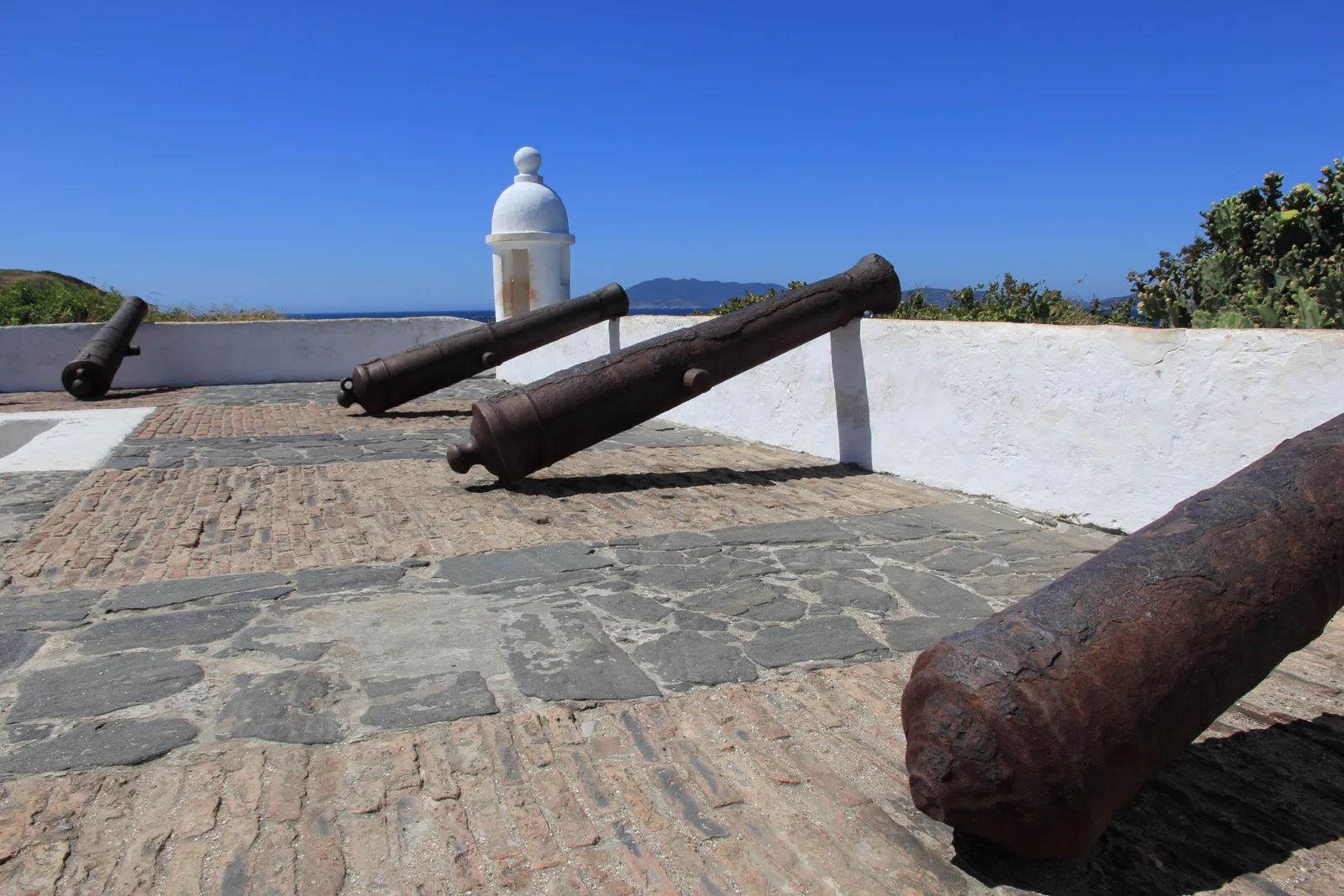 Four old iron cannons resting against the whitewashed parapet on the brick and stone terrace of the Forte São Mateus at Cabo Frio, with a white domed sentry box at the corner and the sea beyond