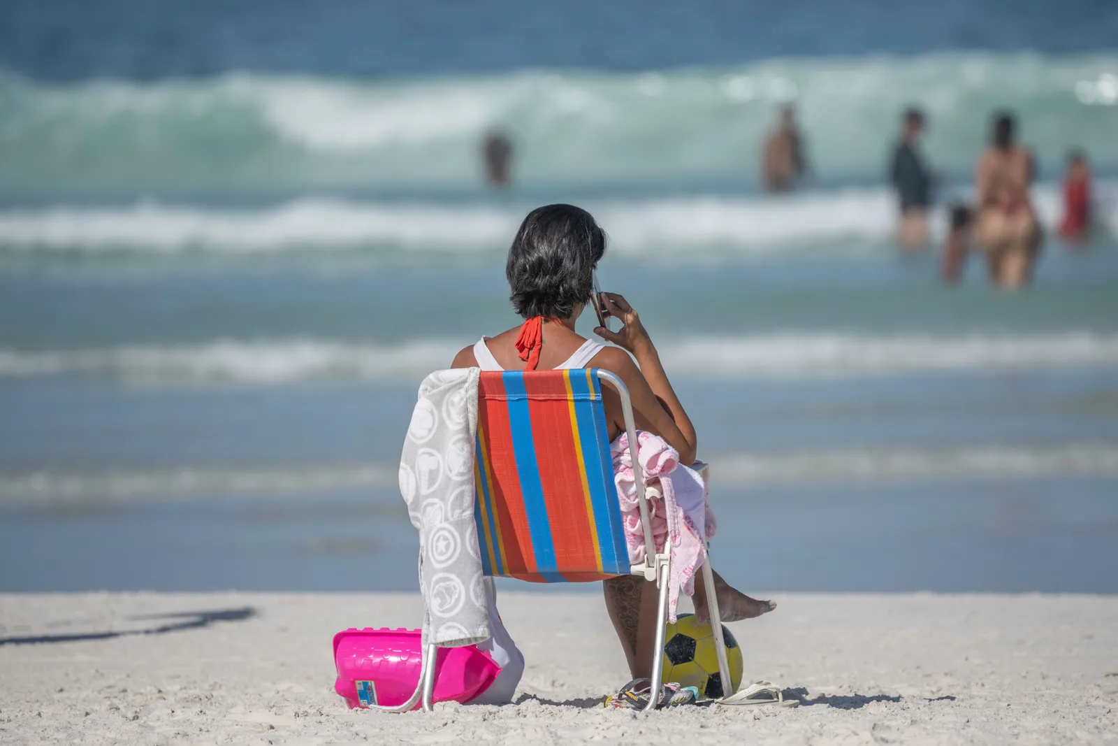 A woman sitting in a striped beach chair on the sand at Cabo Frio with a phone to her ear, surf and other bathers blurred behind her