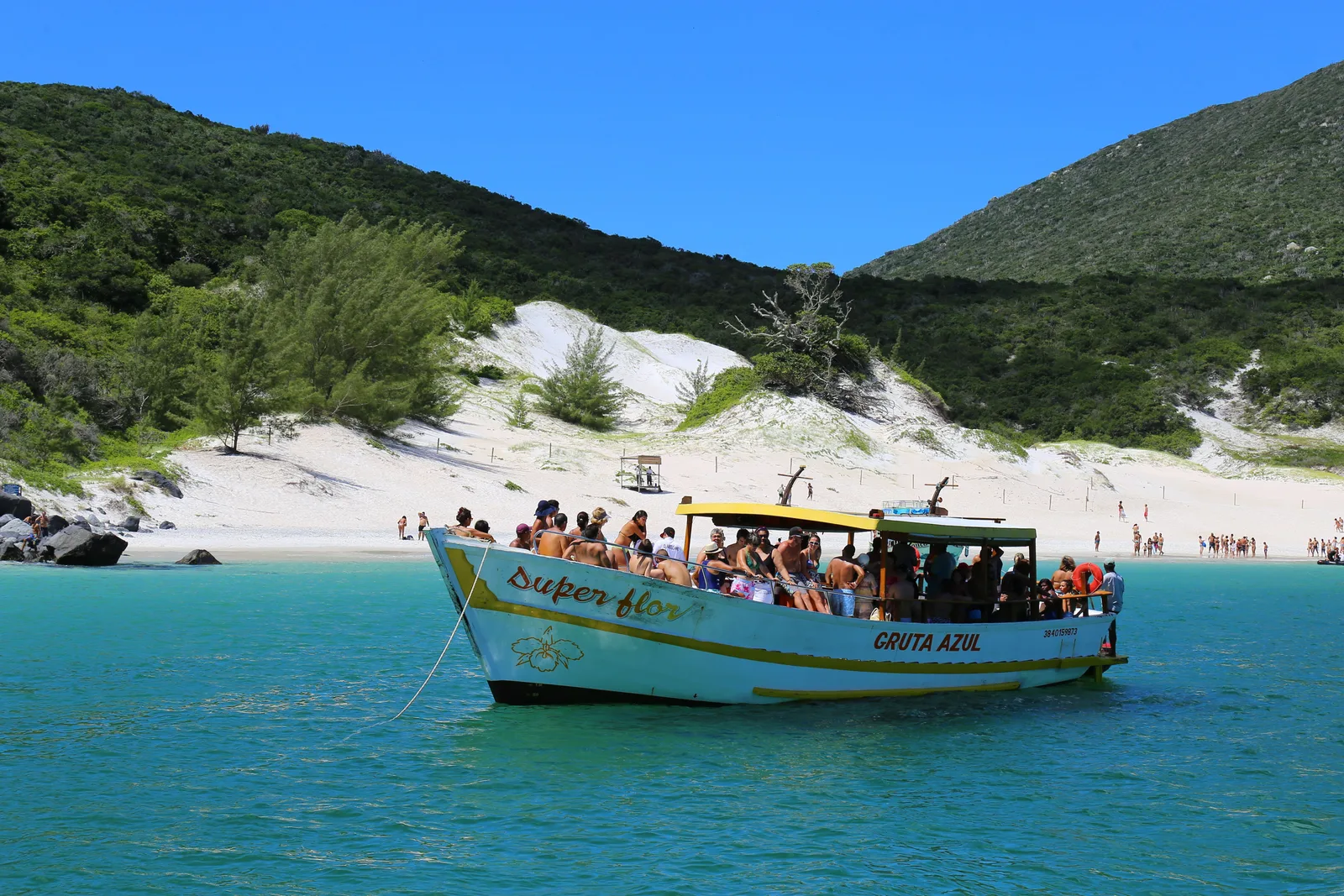 A pale blue wooden tour boat with a yellow and green canopy, crowded with passengers, anchored in turquoise water off a white beach at Arraial do Cabo, with pale dunes and a green hill behind
