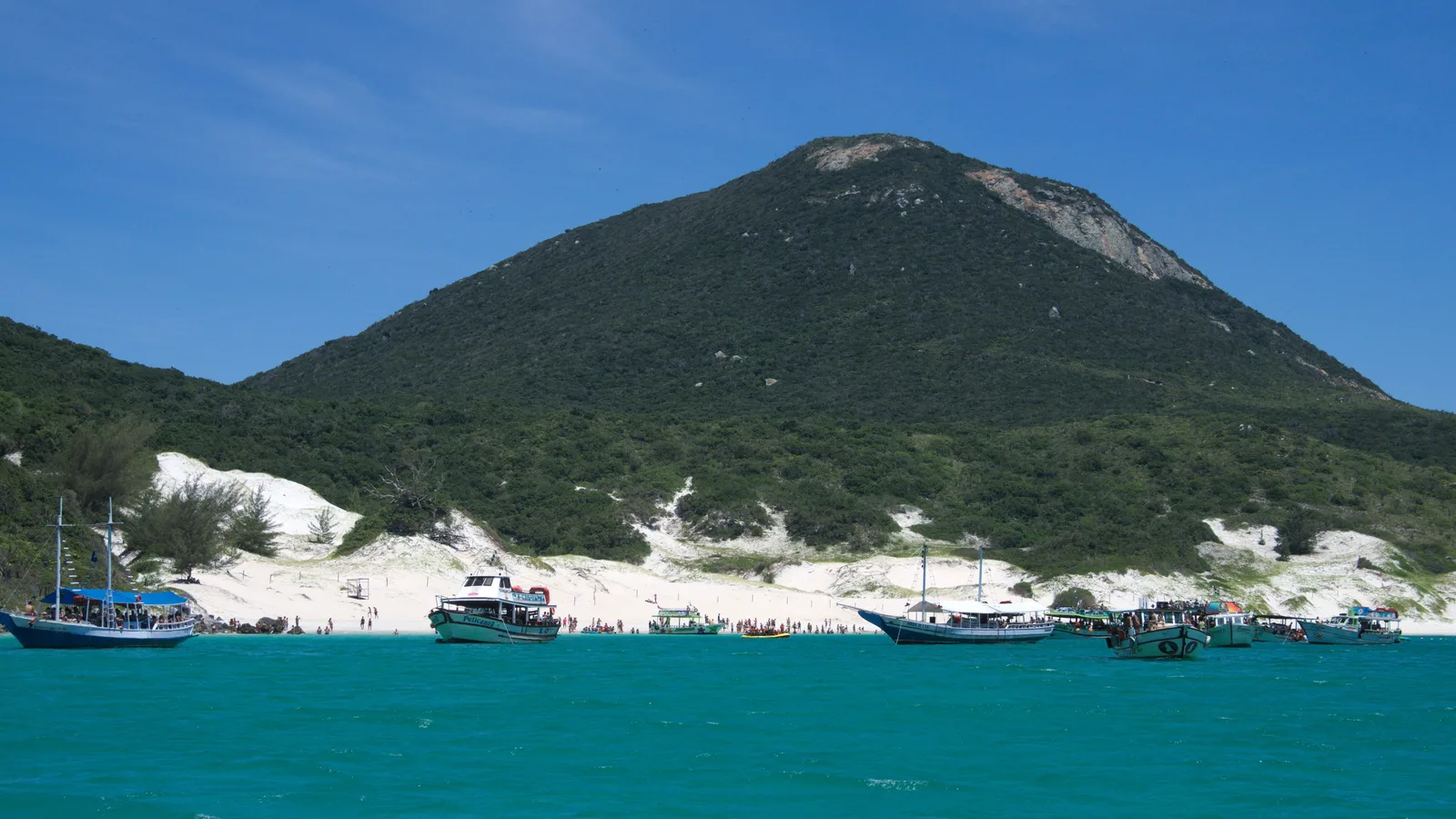 Turquoise water off Praia do Farol at Arraial do Cabo with a line of wooden tour boats anchored close in, a white beach and low white dunes behind them, and the green conical peak of Ilha do Cabo Frio rising above