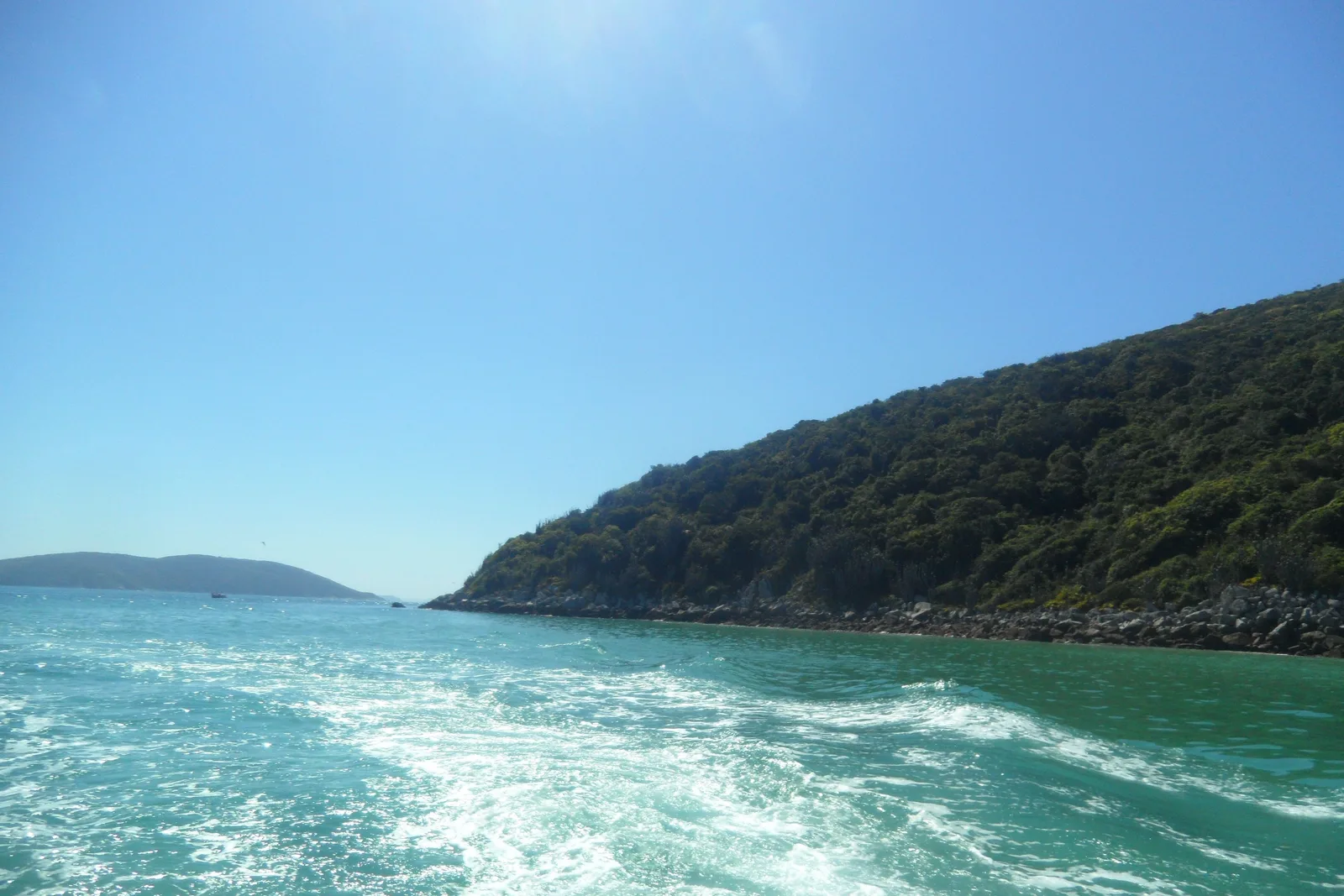 The foaming wake of a moving boat on green-turquoise water at Arraial do Cabo, running past a forested headland with a boulder shore, and a low hill on the horizon