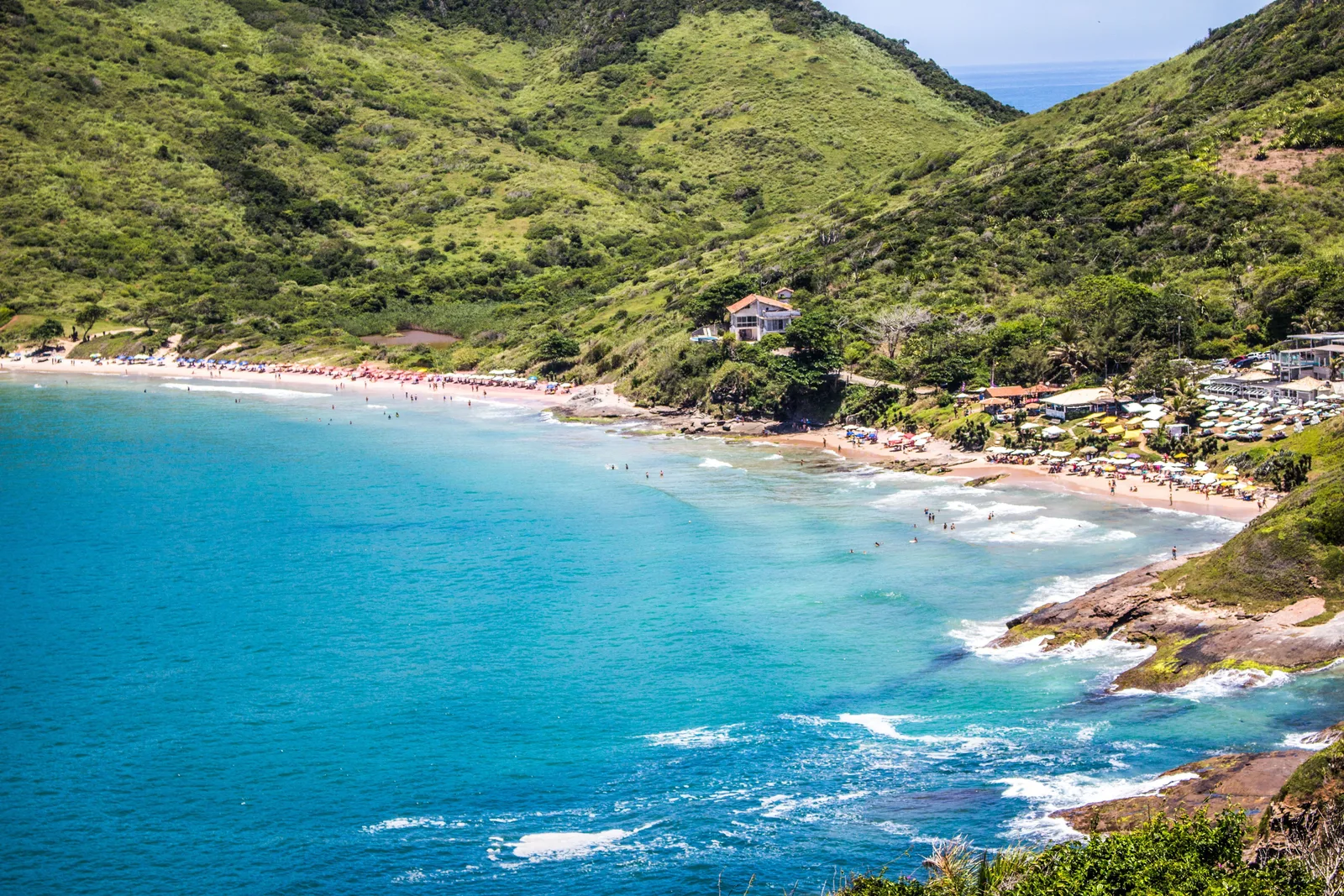 A long curving beach in Búzios lined with parasols and kiosks below steep green hills, with turquoise water and small waves breaking over rocks in the foreground