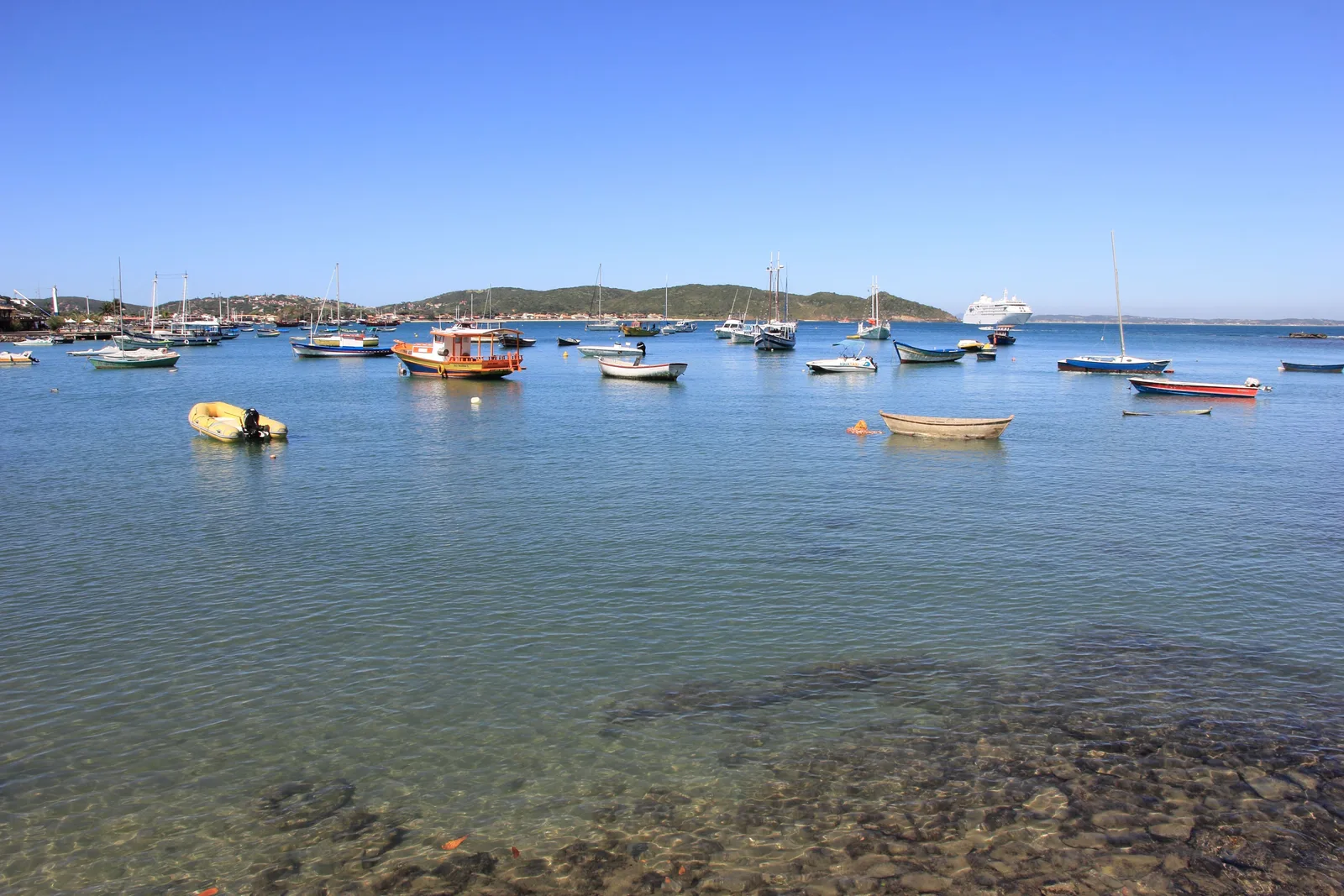 Clear shallow water over a bed of stones in the foreground at Búzios, with dozens of small boats and yachts moored beyond it, a cruise ship out in the bay and low green hills along the far shore