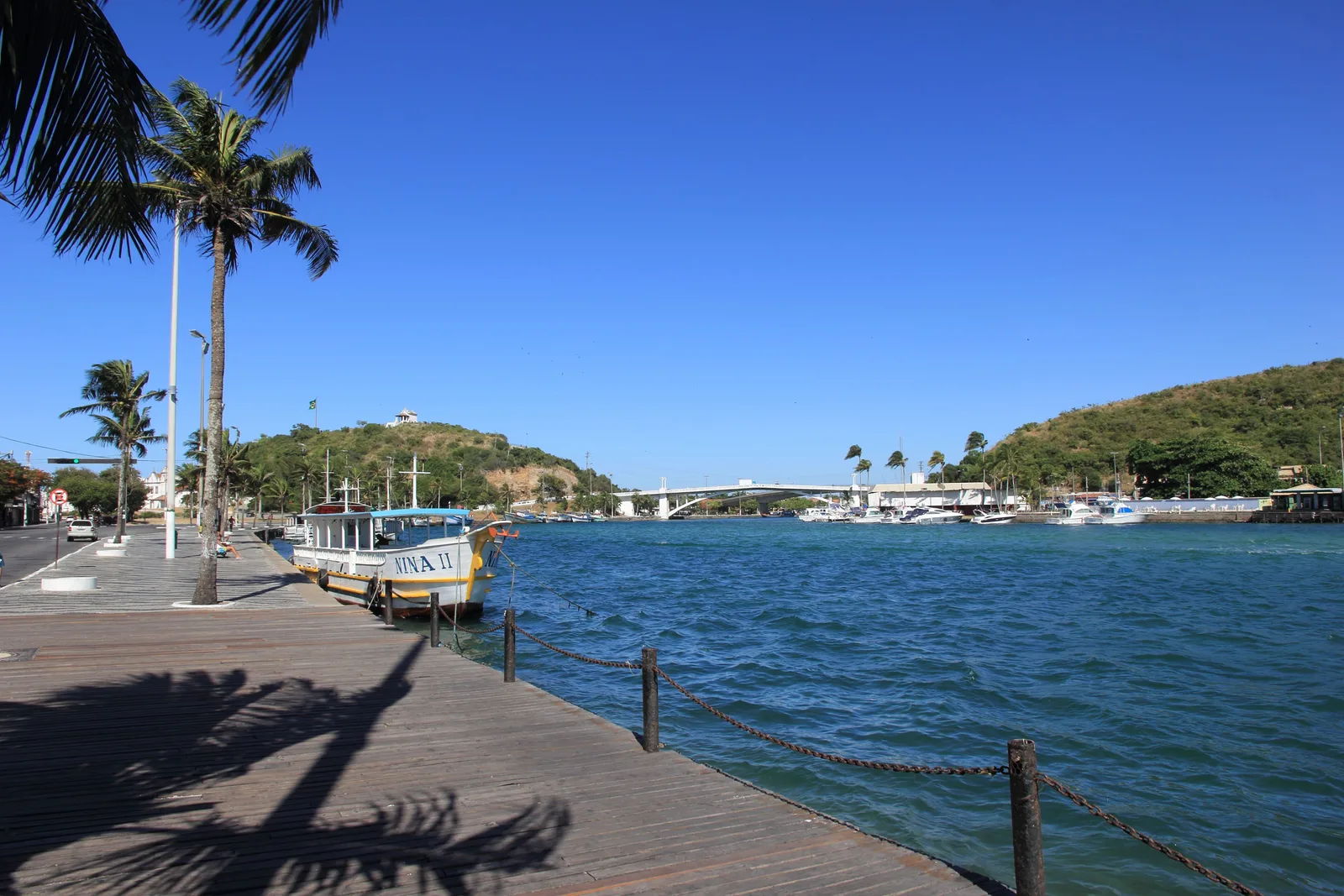 A boardwalk running along the Canal do Itajuru at Cabo Frio under palm trees, with a moored boat on the rippling water and the road bridge crossing the channel beyond