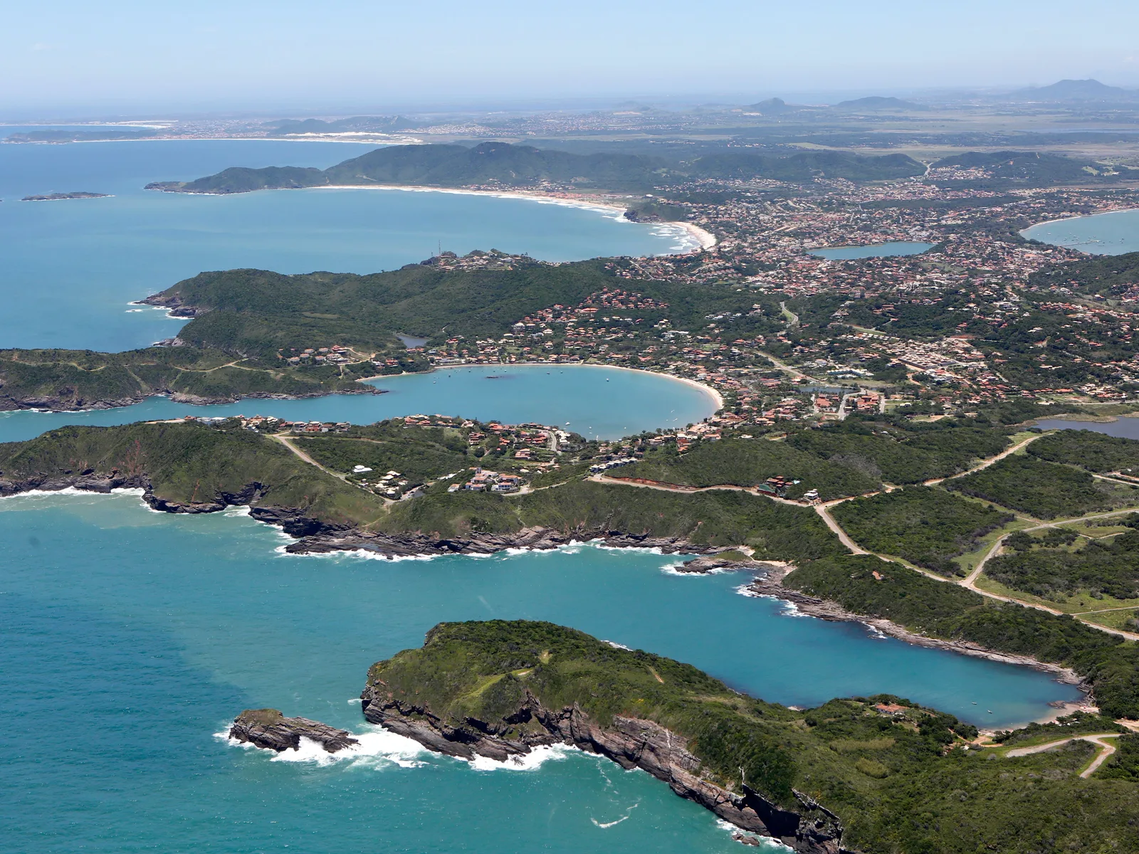 The Búzios peninsula from the air: a headland of low green hills with several separate curving bays cut into it, pale sand along each one and the town spreading inland behind