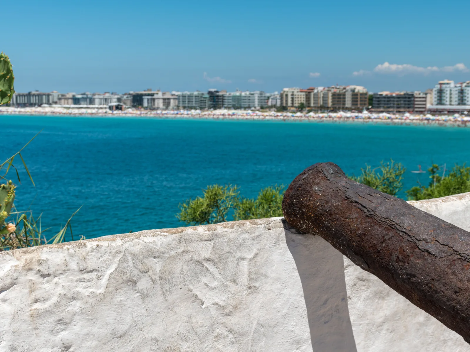 An iron cannon on the whitewashed rampart of the Forte São Mateus at Cabo Frio, pointing out over turquoise water toward the long city beach and the line of apartment towers behind it