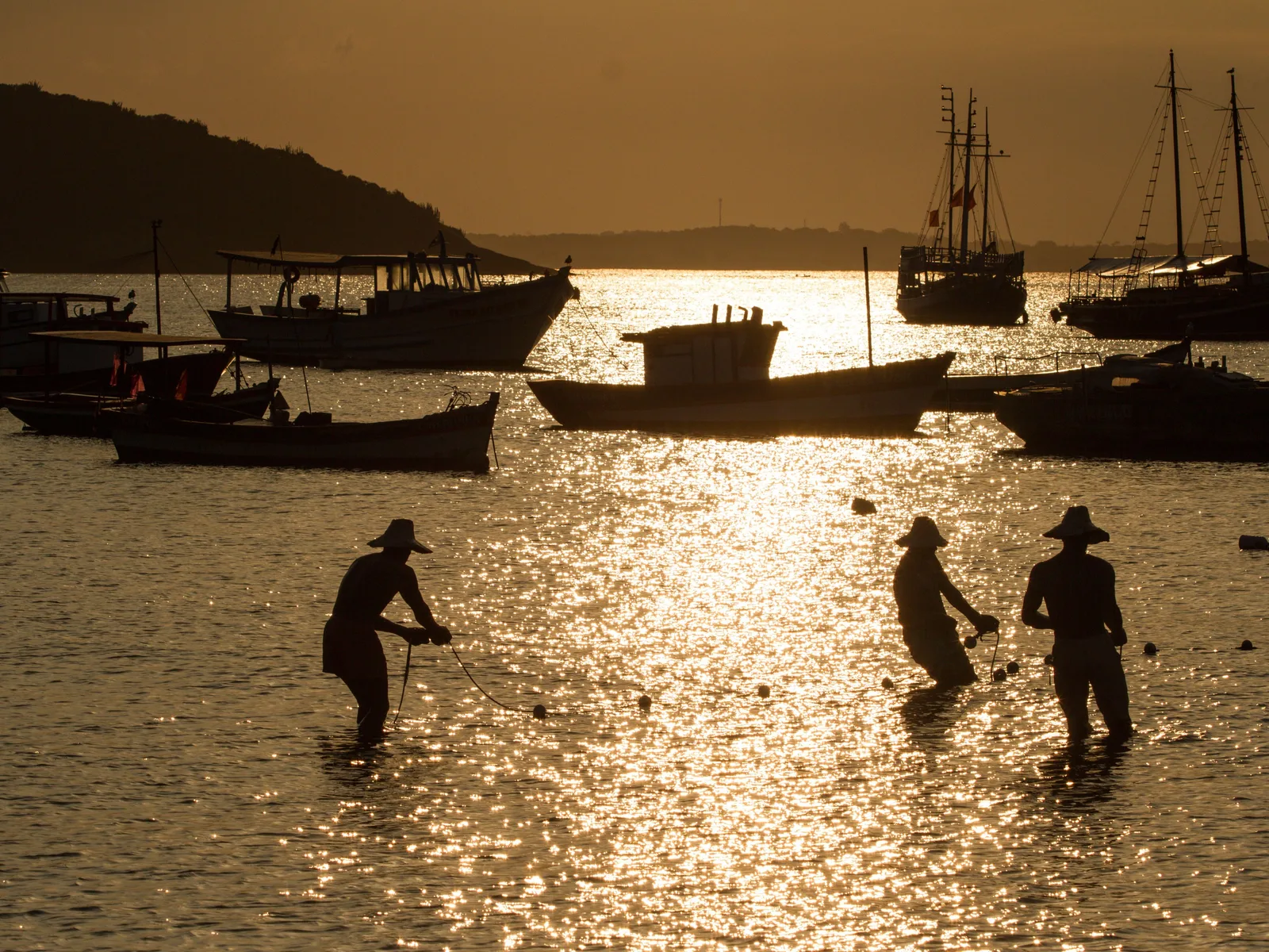The Monumento dos Três Pescadores on the Orla Bardot at Búzios at sunset: three bronze fishermen silhouetted standing in shallow gold water hauling a net, with moored boats and masts behind them