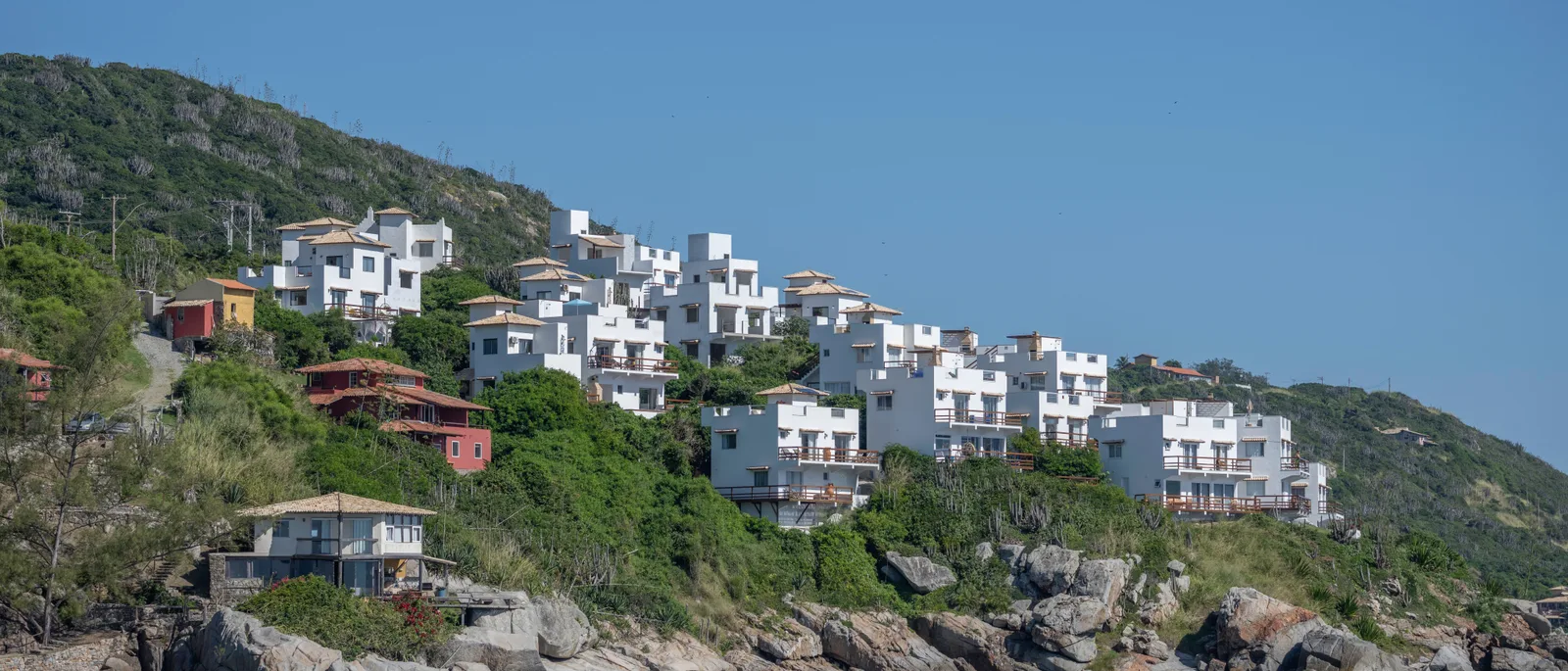 White houses and a few red ones stacked up a steep green hillside at Arraial do Cabo in a wide frame, with gray boulders along the foot of the slope