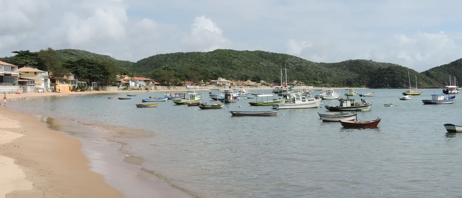 The waterfront at Praia da Armação in Búzios in a wide frame: a curve of sand with small boats moored just offshore, low buildings along the back of the beach and green hills behind the town