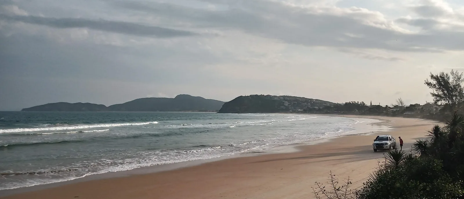 Praia de Geribá in Búzios in a wide frame under a cloudy sky: a long curve of sand with small surf breaking, a pickup truck parked on the beach, a low headland with houses at the far end and palms in the foreground