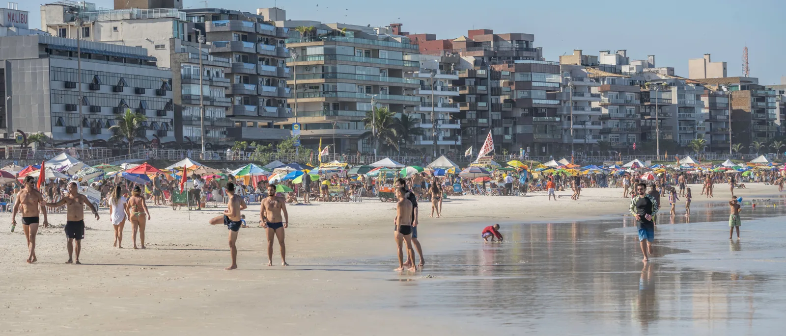 A wide frame of the beach at Cabo Frio: rows of colored parasols packed along the back of the sand, people walking on the wet foreshore, and a solid wall of tall apartment blocks behind