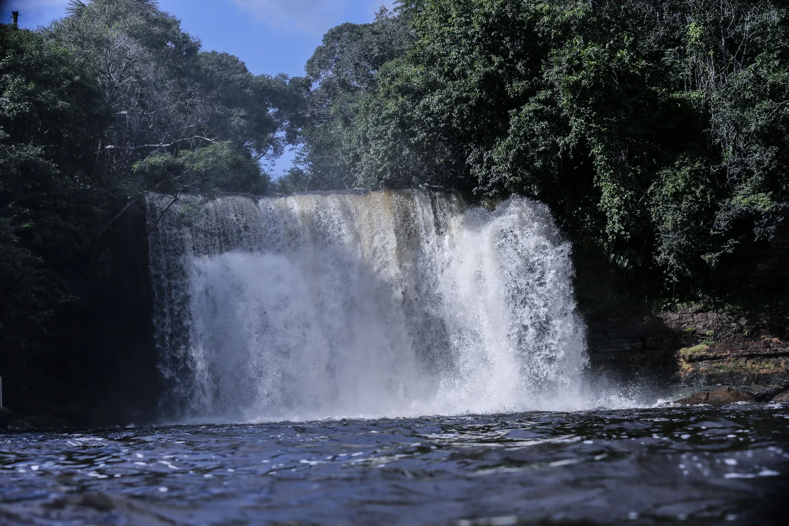 A broad waterfall spilling off a rock shelf into dark water, dense forest on both sides and a strip of blue sky above