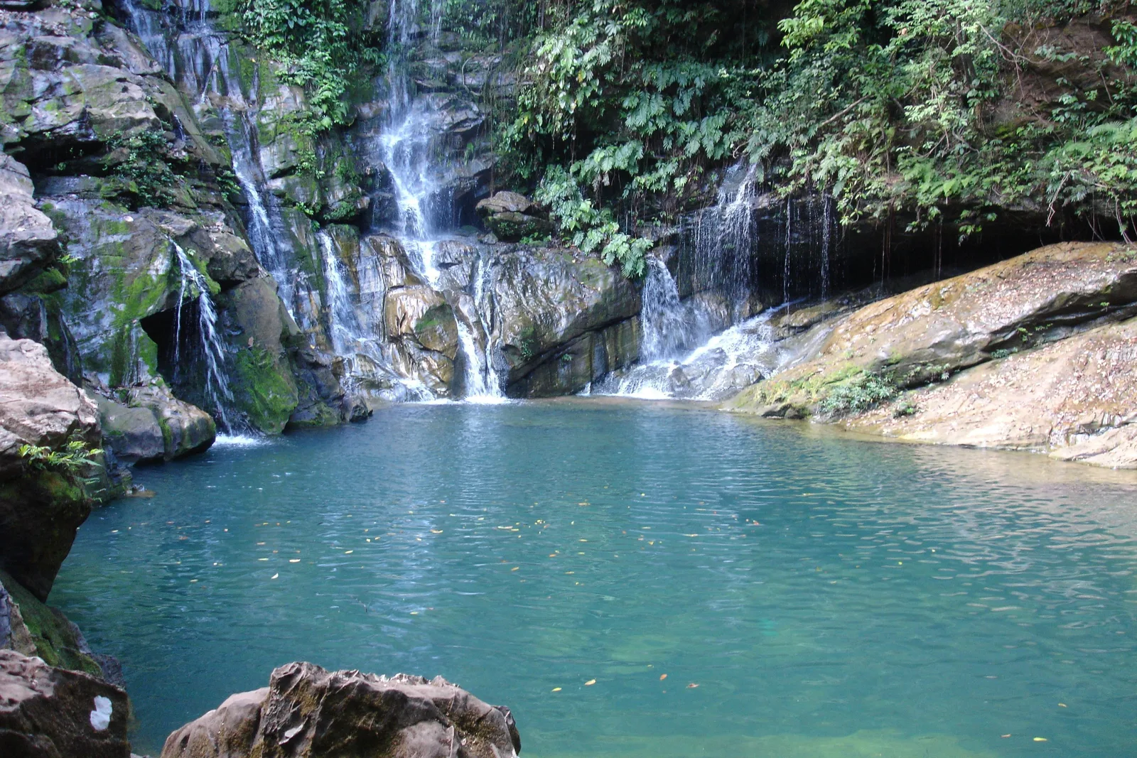 Thin streams of water falling down mossy gray rock ledges into a still turquoise pool edged with boulders