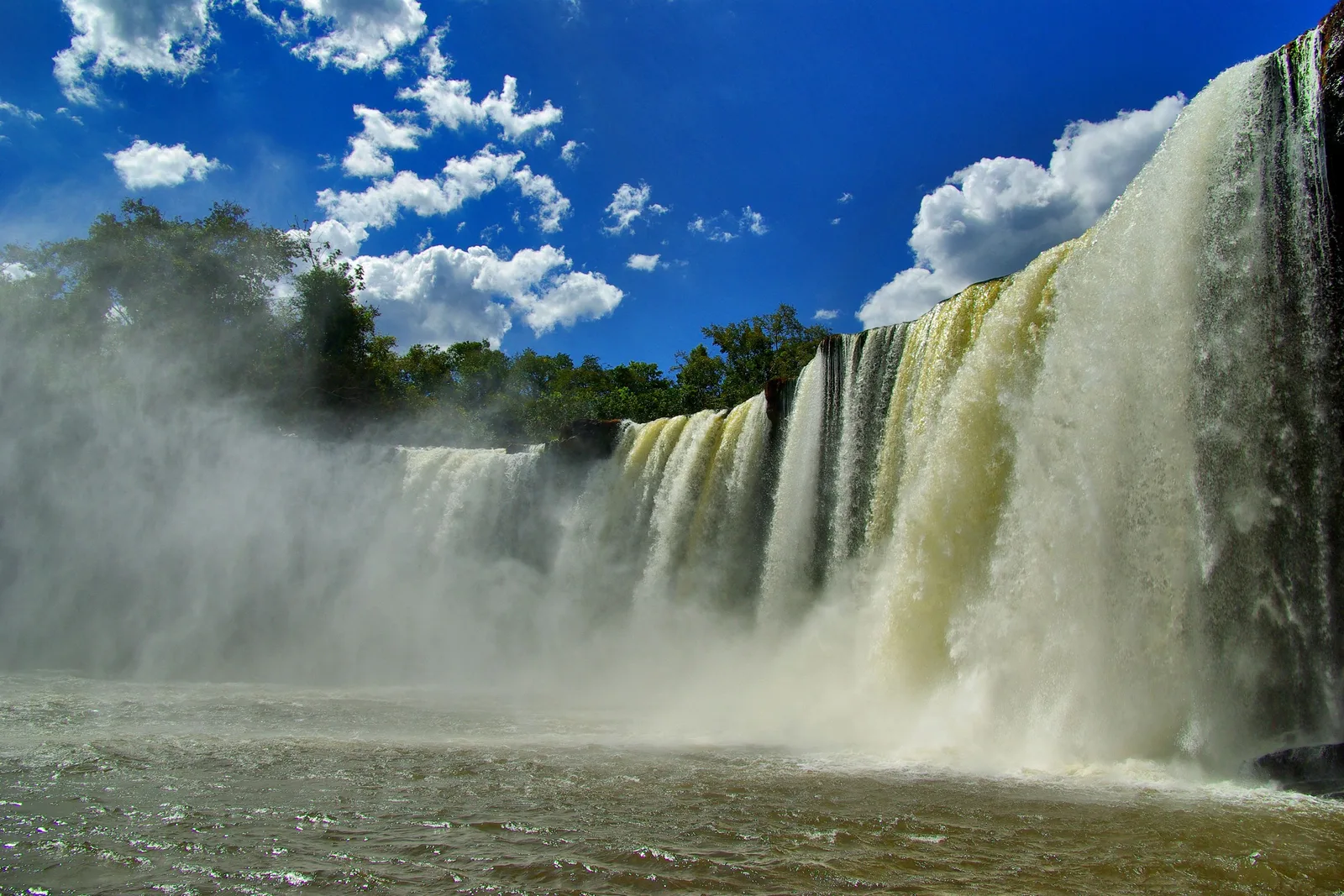 A broad waterfall seen from the pool below, white water and mist across the whole frame under a deep blue sky