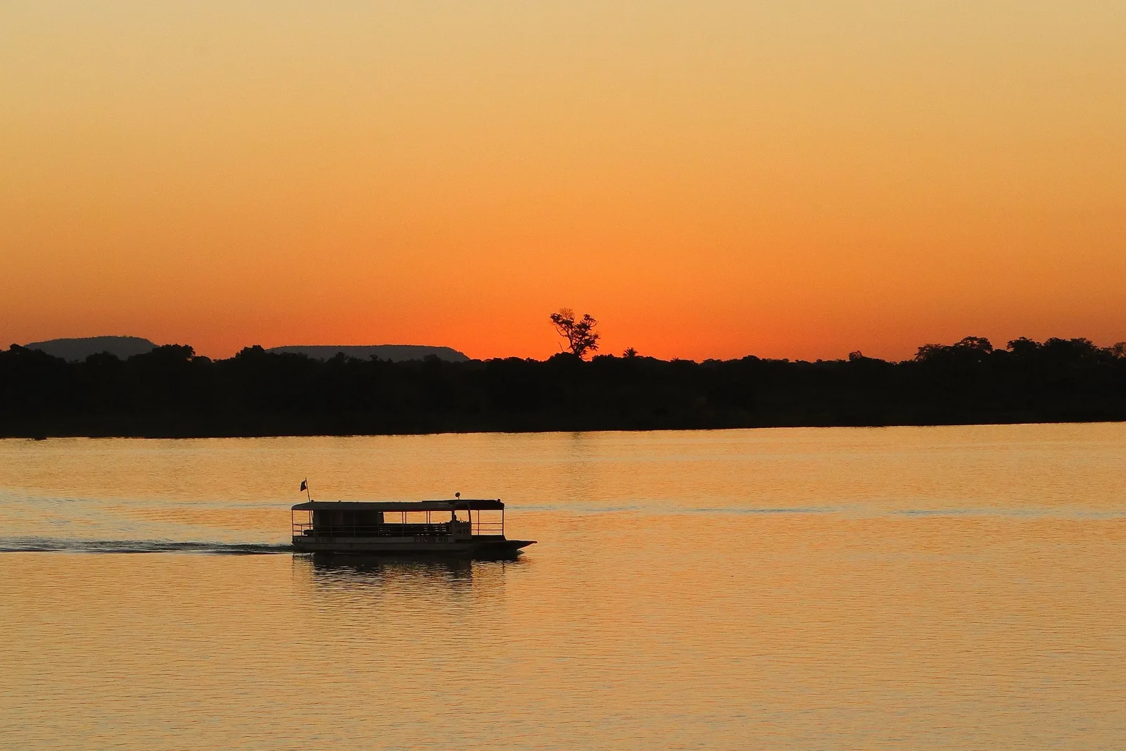 A passenger boat crossing a wide river under an orange sunset sky, the far bank a dark line of trees