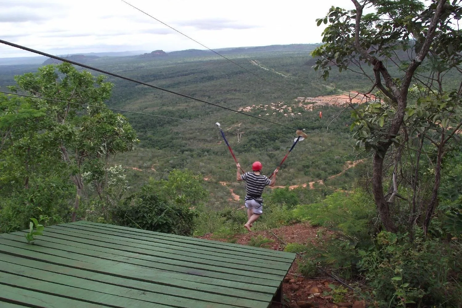 A rider in a harness gliding away down a zipline cable over a wooded valley, seen from a wooden launch platform