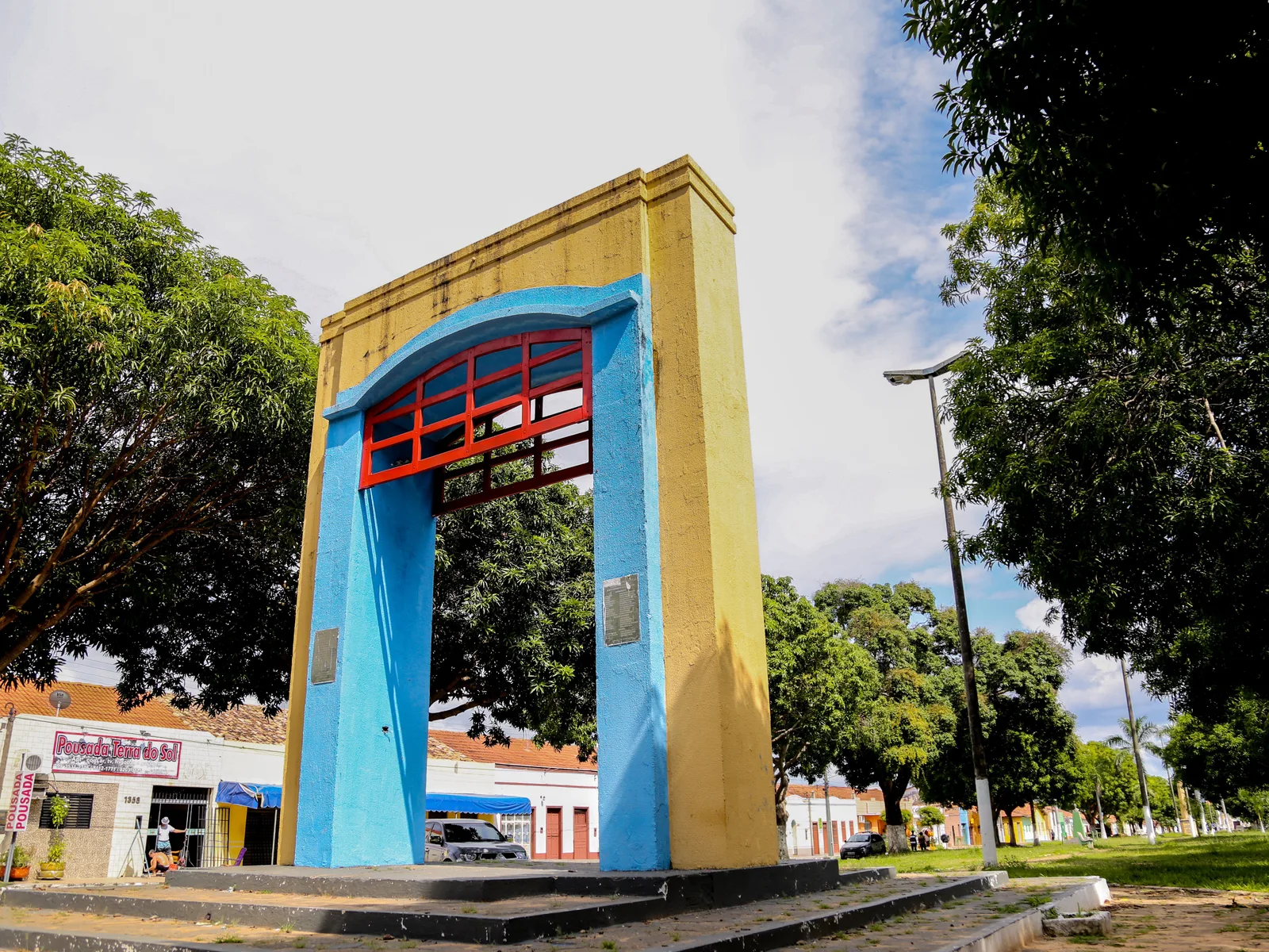 A tall yellow monument shaped like a gateway, with a blue arch and red lattice inside it, standing on a square in Carolina among trees and low shops