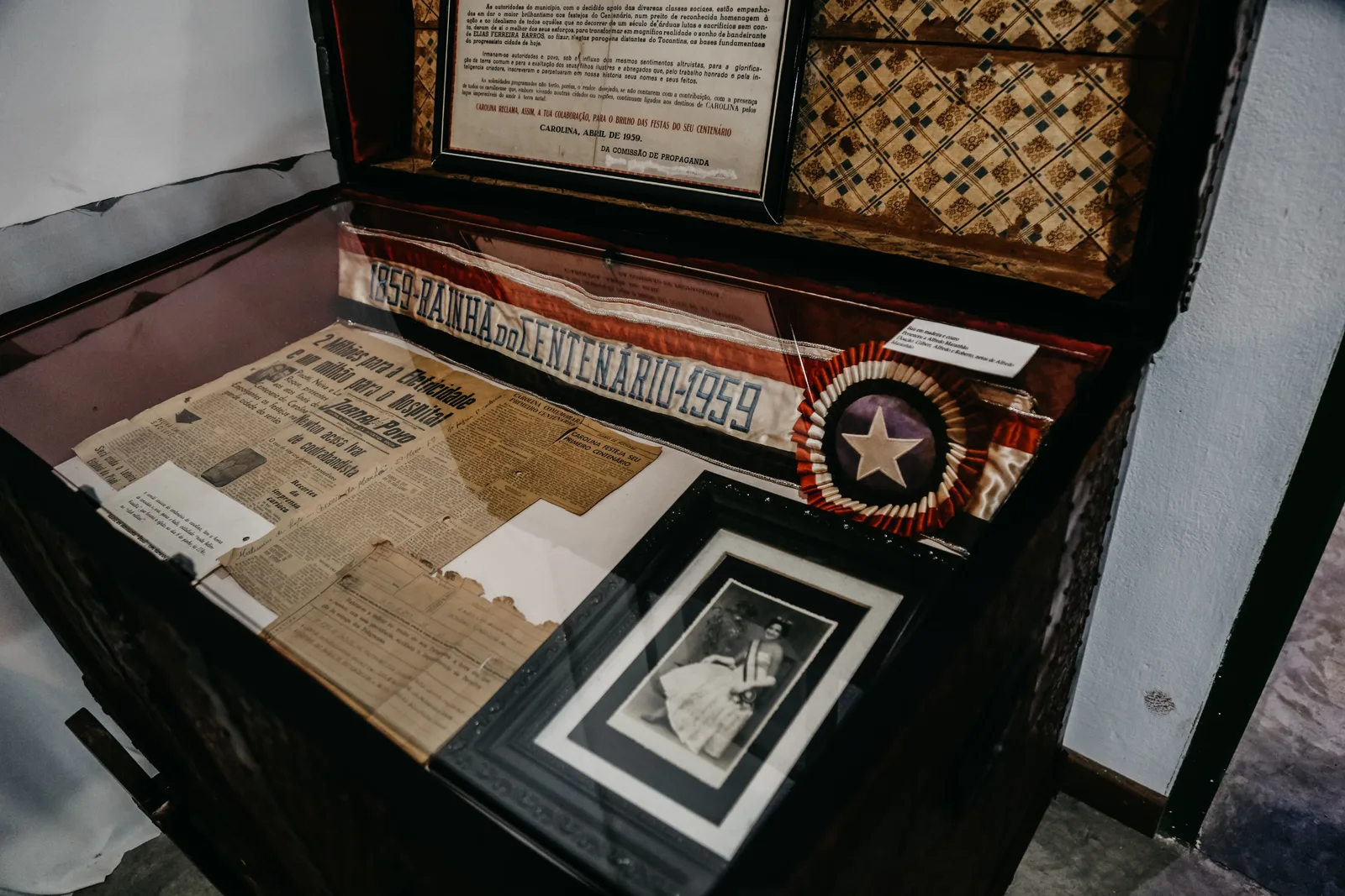 A glass display case in the Museu Histórico de Carolina holding old newspapers, a framed portrait and a ribbon with a star