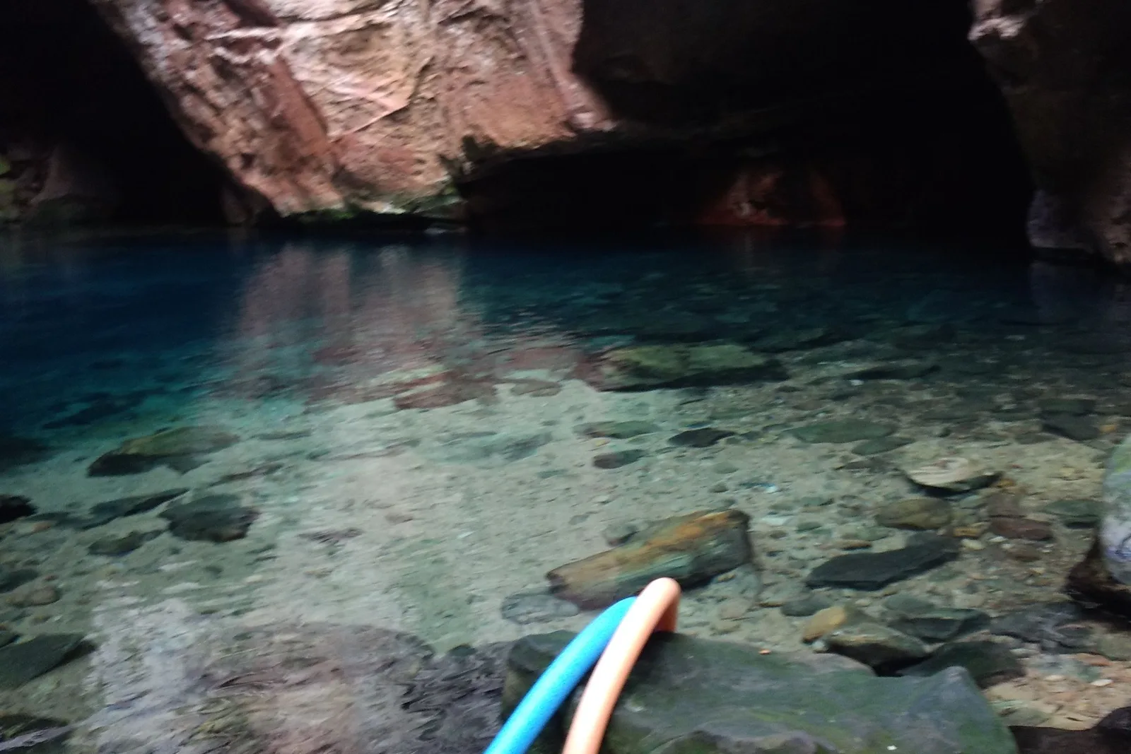 Clear blue-green water over a pebbled bottom beneath an overhanging rock face, the ends of two foam pool noodles, blue and peach, in the foreground