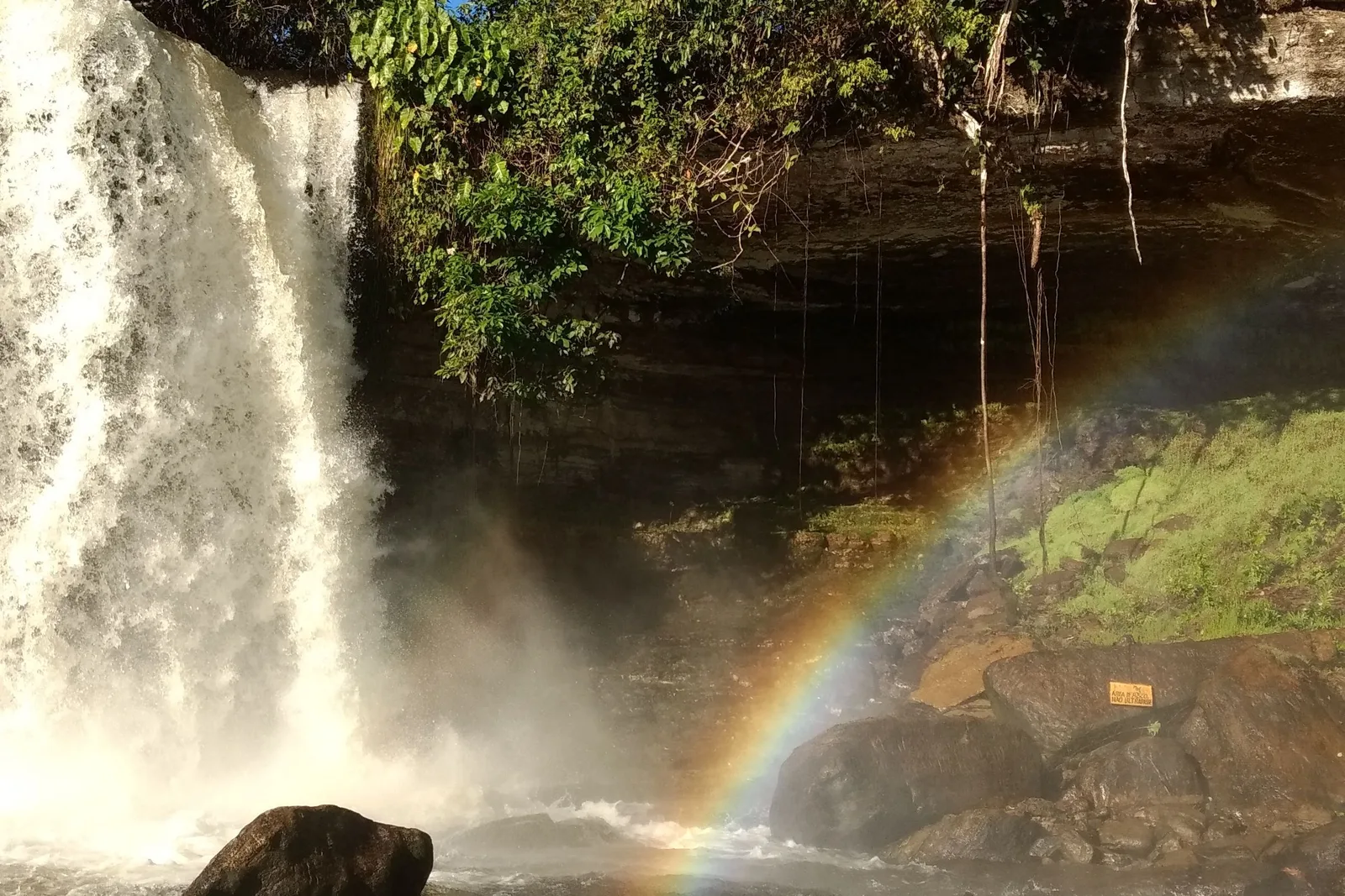 A waterfall pouring off a rock ledge into a dark river with a rainbow arcing across the spray