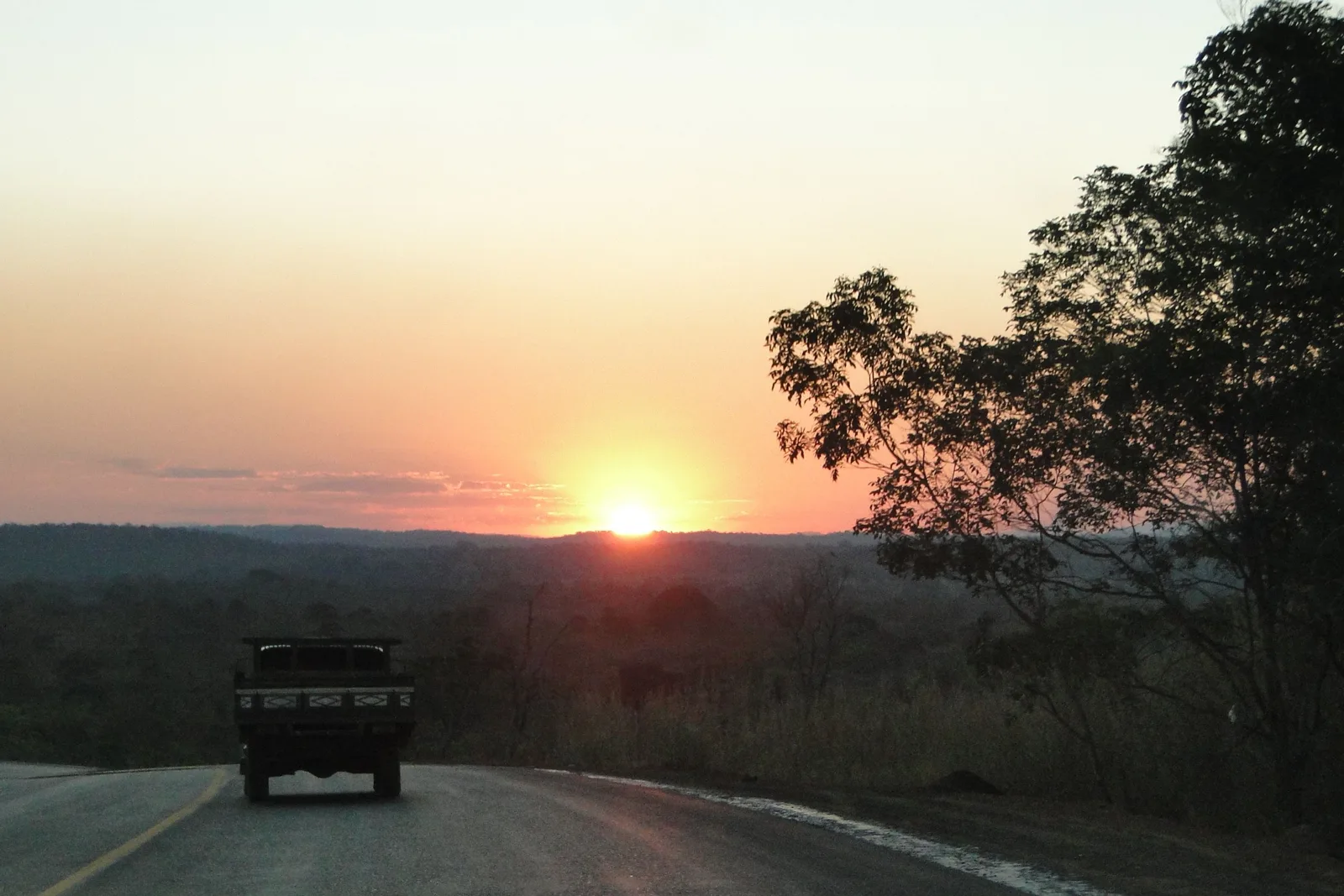 A pickup truck driving down an empty highway toward a low sun setting over a wide wooded plain