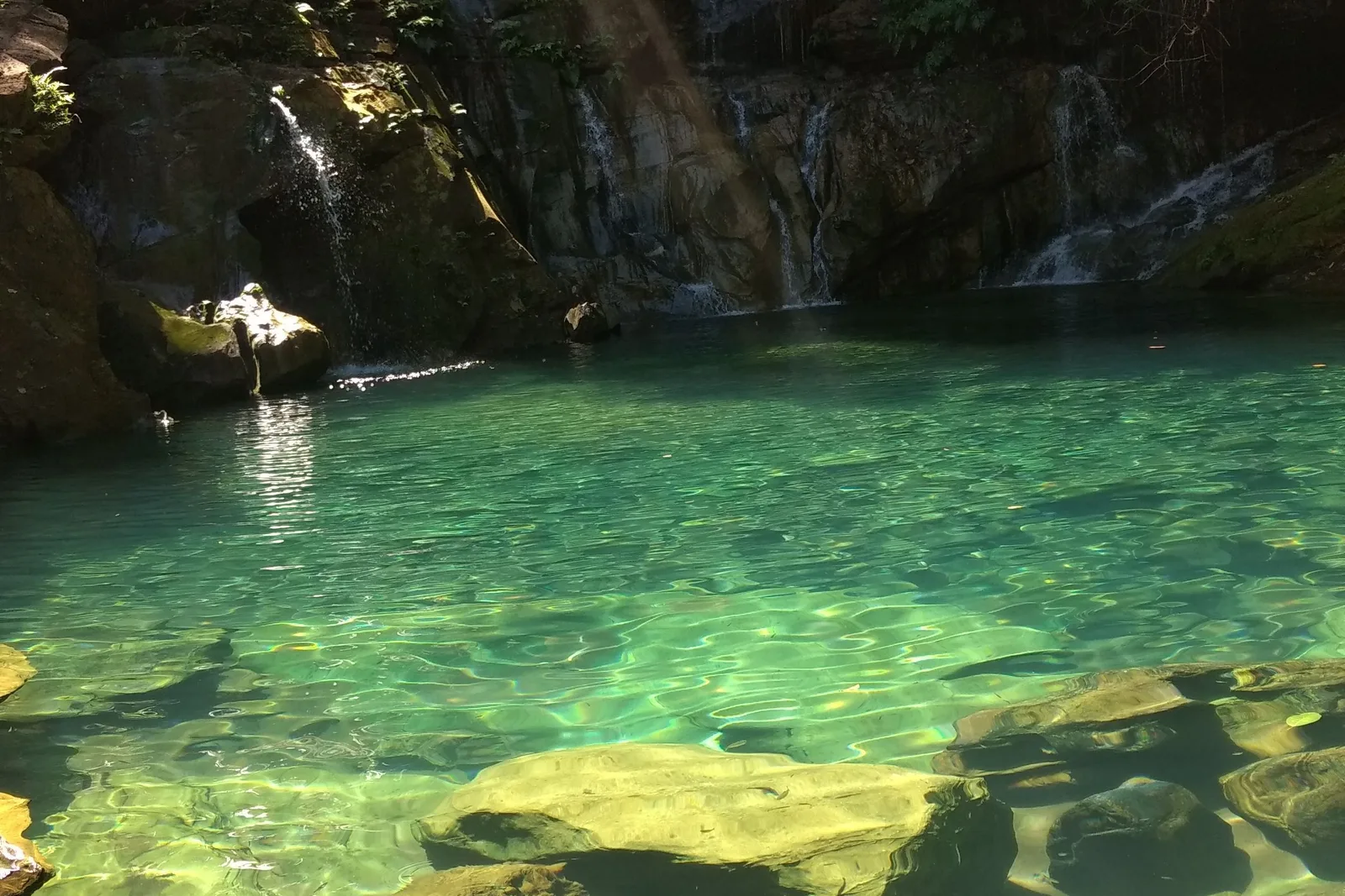 A clear green pool with sunlit boulders under the surface in front and thin waterfalls running down the dark rock wall behind
