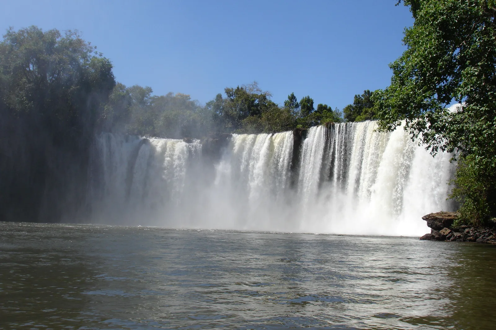 A wide waterfall falling in a long white curtain into a dark pool, with forest above and a rock on the right bank