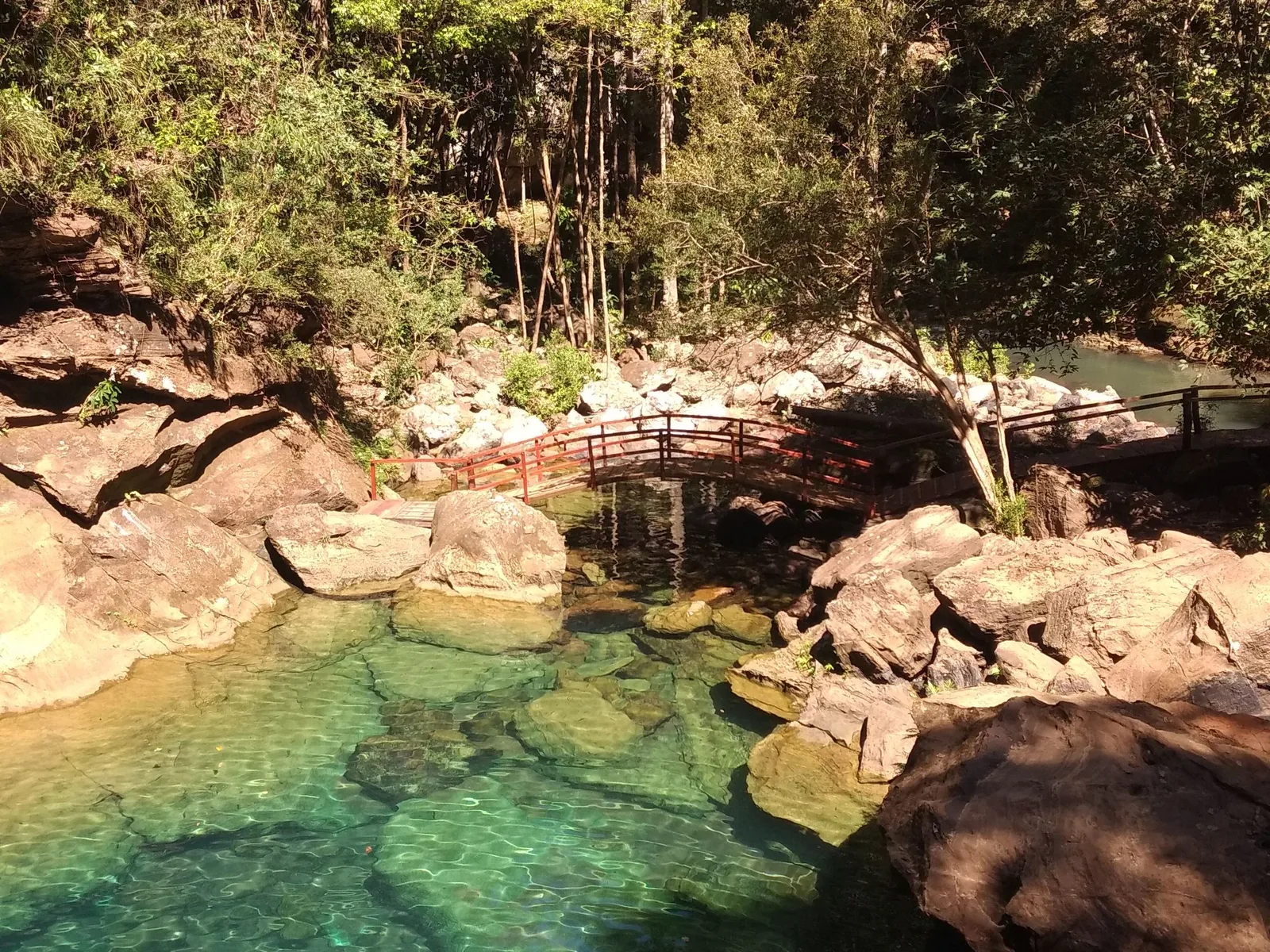 A clear green pool among pale boulders in a wooded gorge, a wooden footbridge crossing the rocks behind it